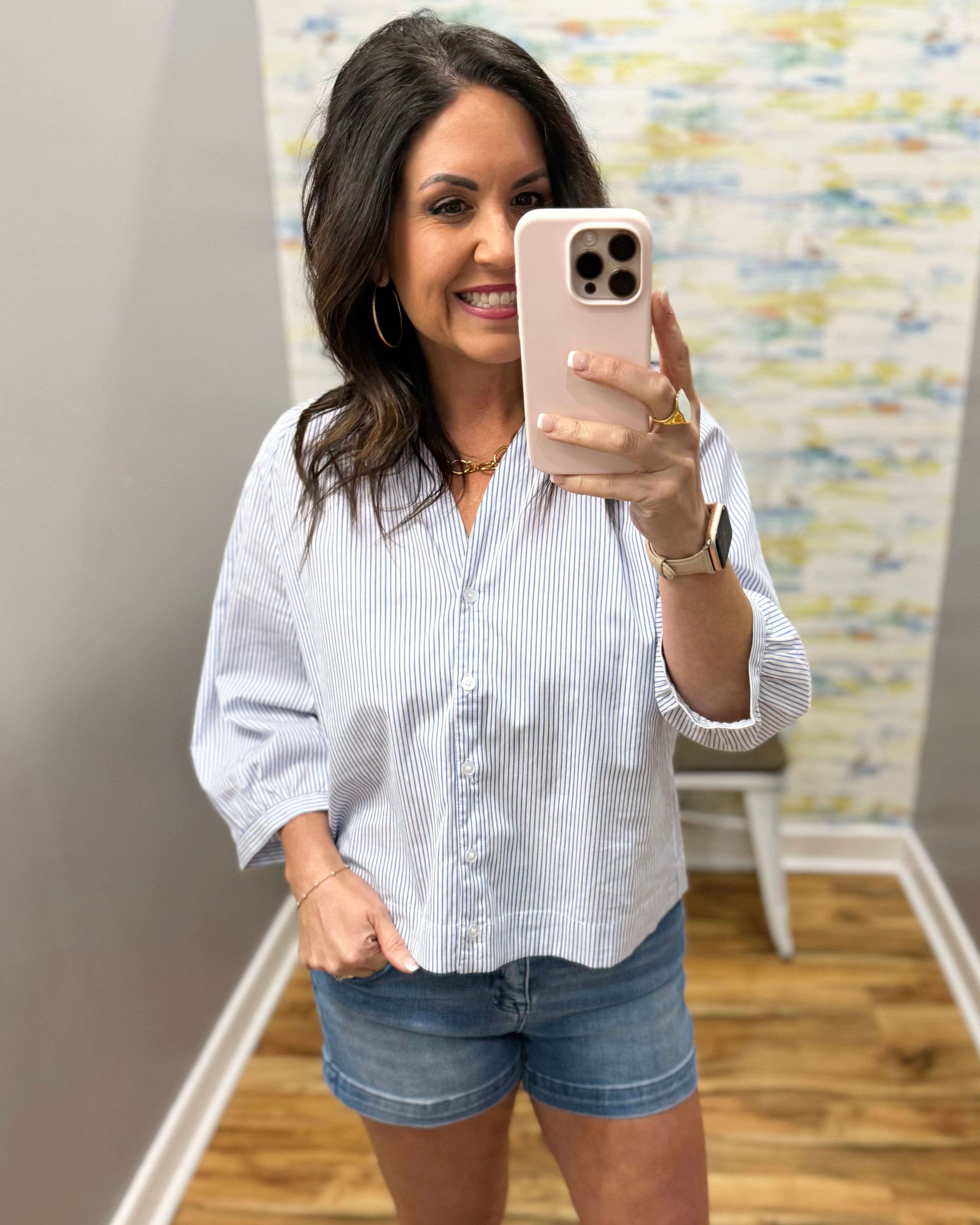 Woman taking a mirror selfie wearing a white striped shirt and denim shorts.