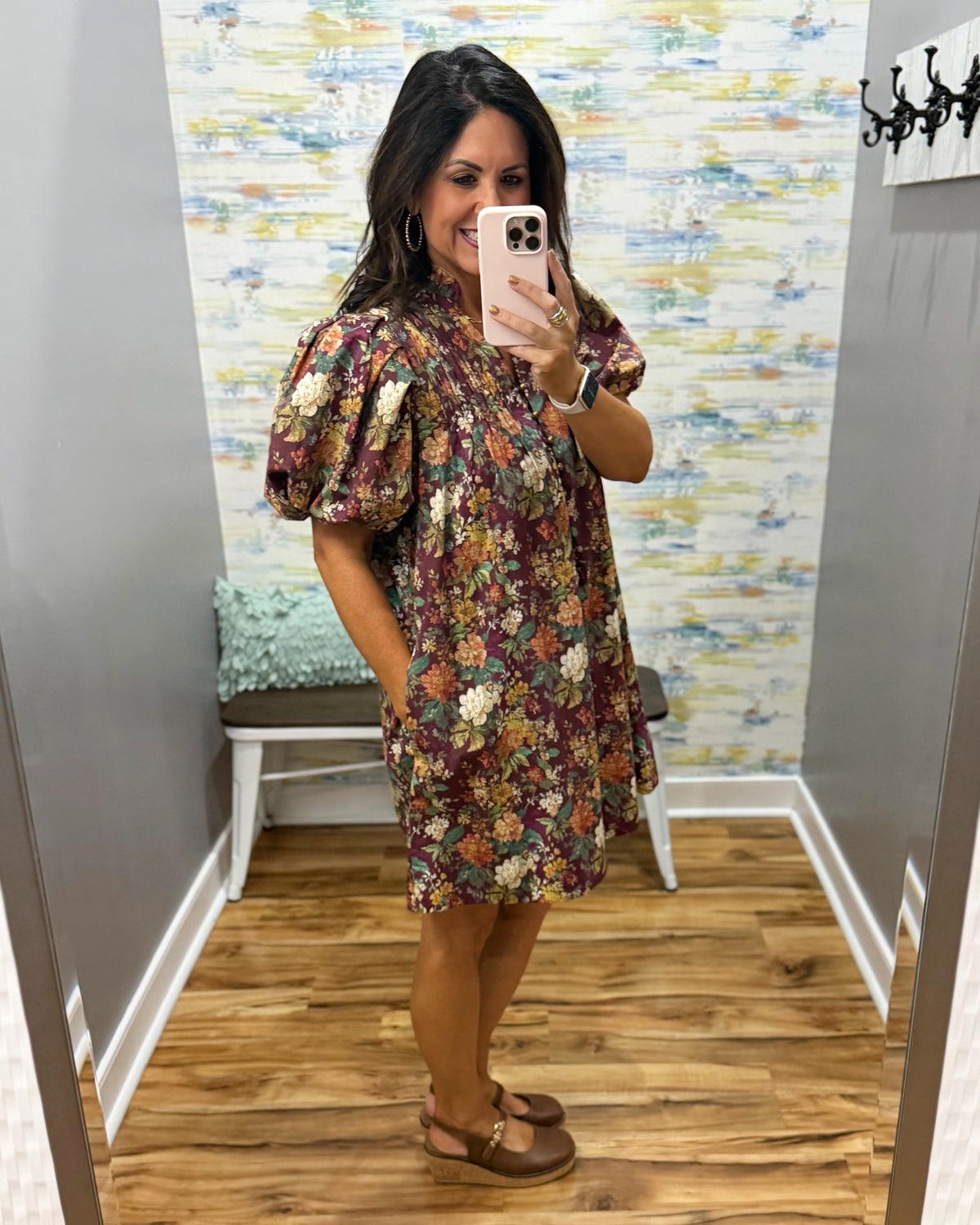 A woman takes a mirror selfie in a fitting room wearing a short puff-sleeve dress with an all-over floral print in earthy tones of brown, burgundy, green, and cream. She is wearing brown flat sandals and holding a light pink smartphone.