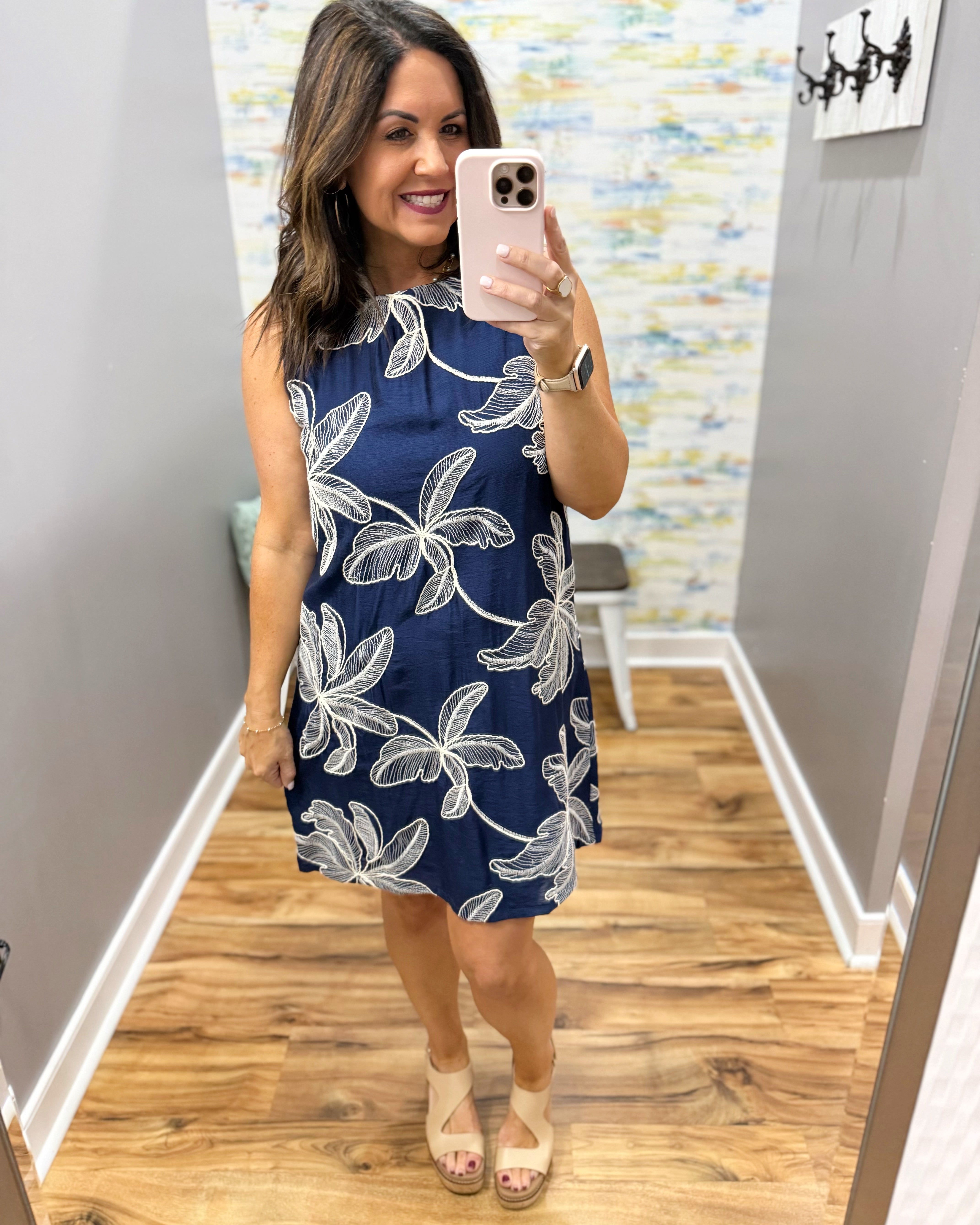Woman taking a mirror selfie wearing a navy dress with white floral patterns in a room with wooden flooring and a colorful wall.