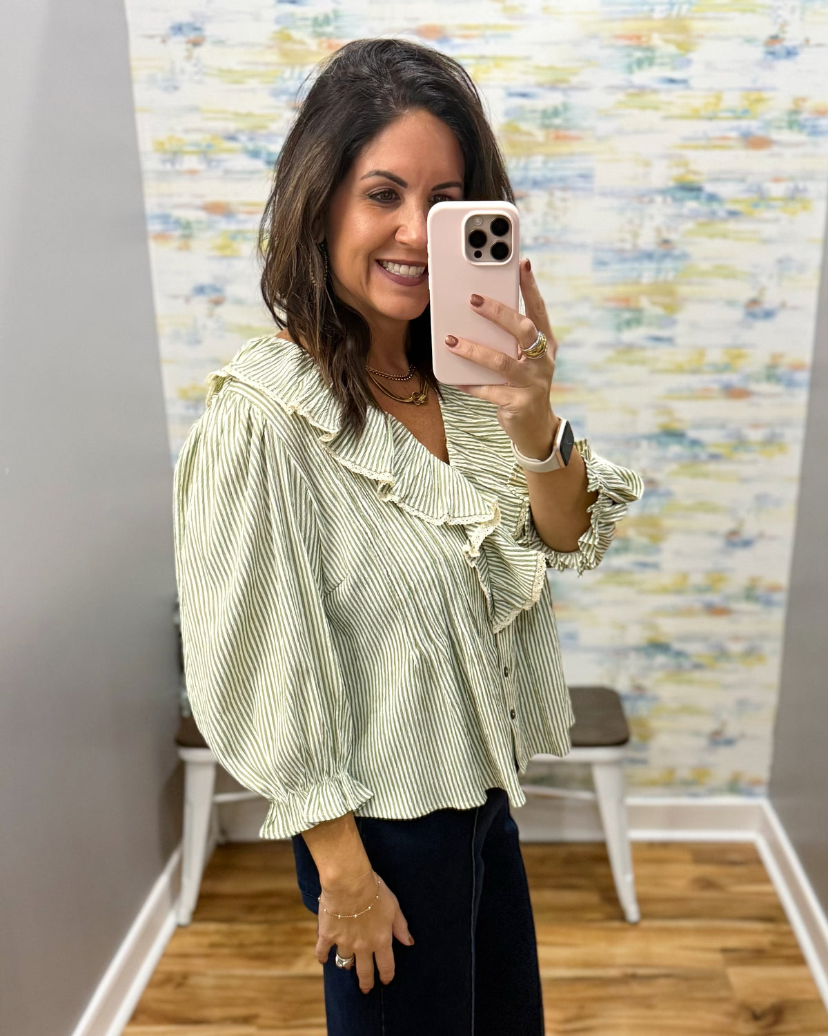 A woman takes a mirror selfie wearing a light green and white vertically striped blouse with ruffle details on the collar and sleeves, paired with dark pants.
