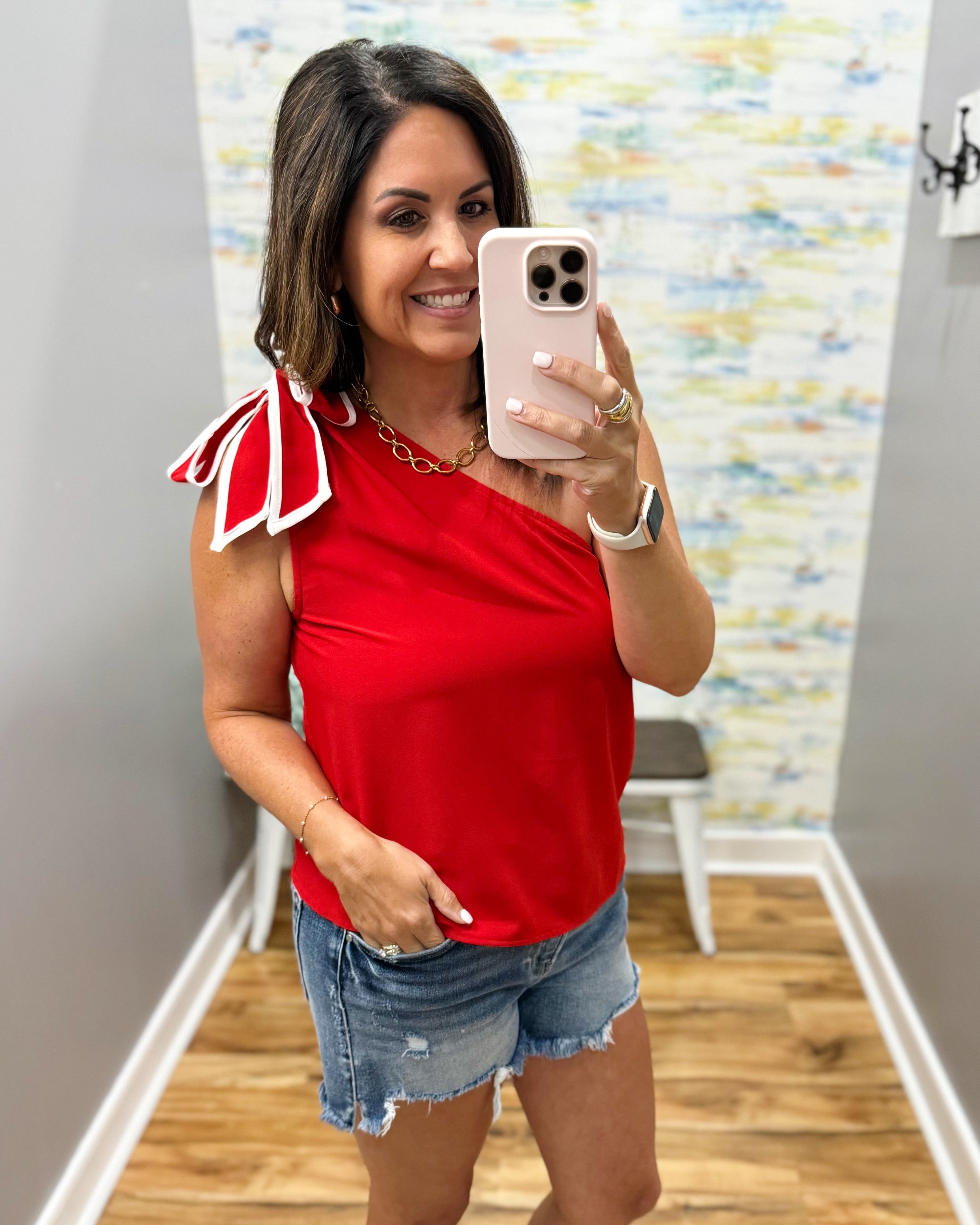 Woman taking a mirror selfie wearing a red top with bow details and denim shorts.