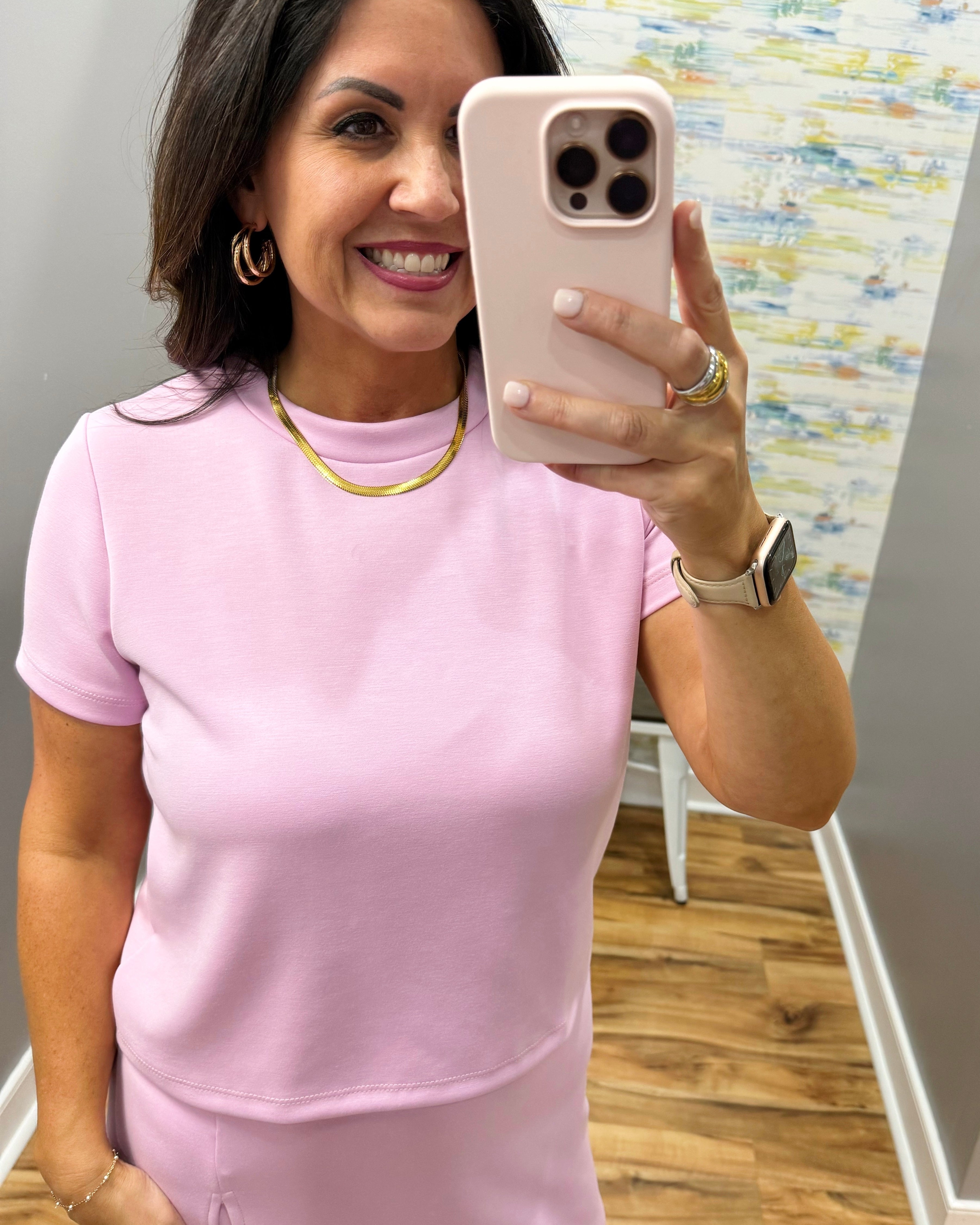 Woman taking a mirror selfie wearing a pink outfit with a colorful wall in the background.