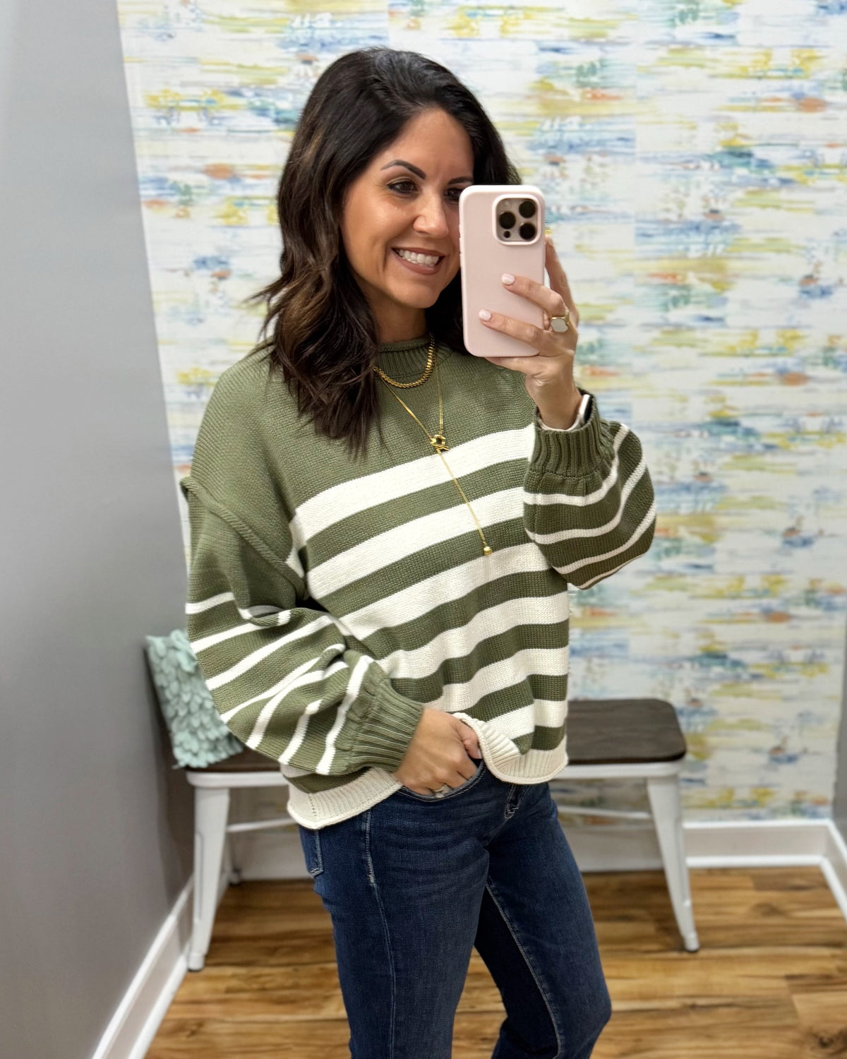 A woman takes a mirror selfie wearing an olive green and cream striped knit sweater, blue jeans, and gold necklaces in what appears to be a boutique fitting room.