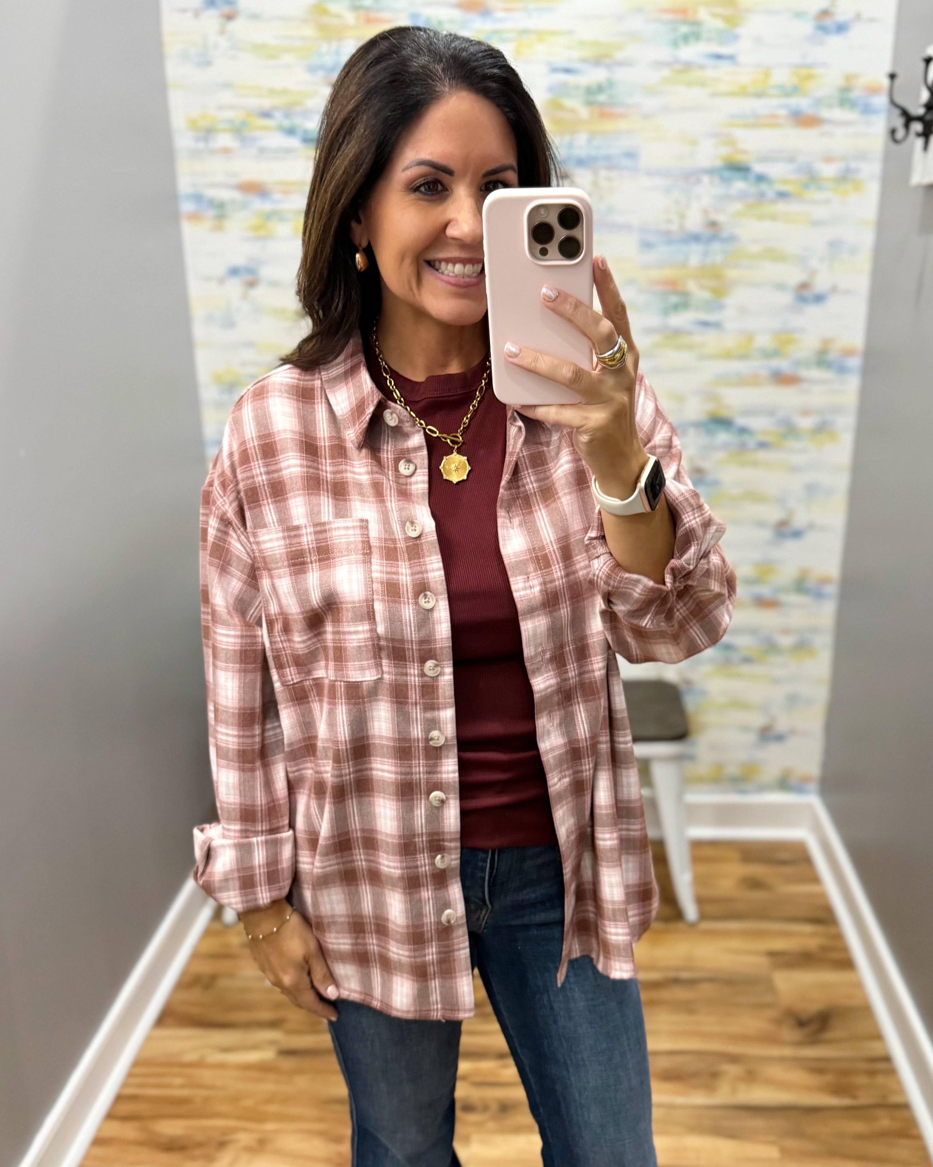 A woman takes a mirror selfie wearing a pink and white plaid button-down shirt over a burgundy top and blue jeans.