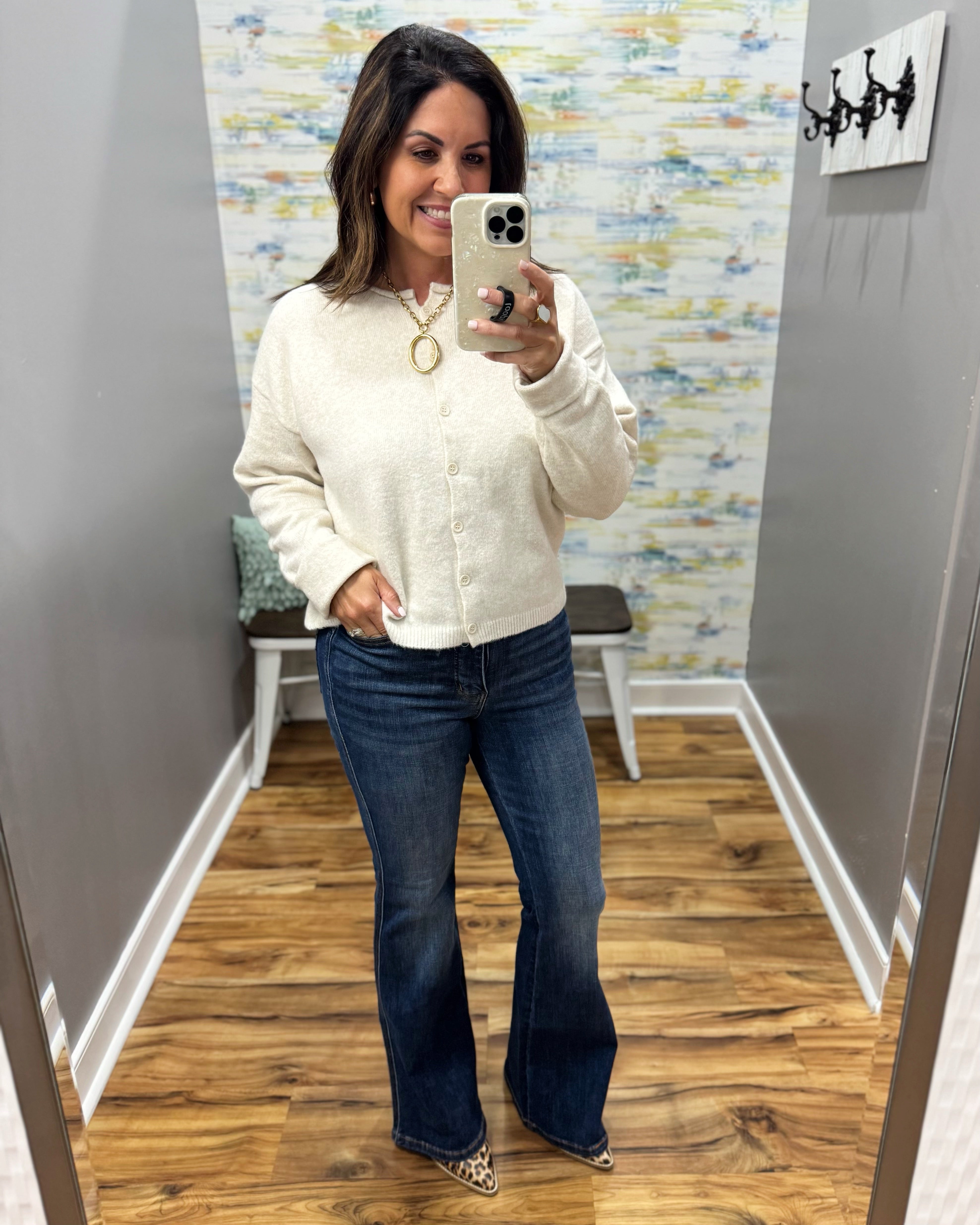 A woman takes a mirror selfie in a fitting room wearing a cream-colored textured knit cardigan, dark blue flare jeans, and leopard print heels.