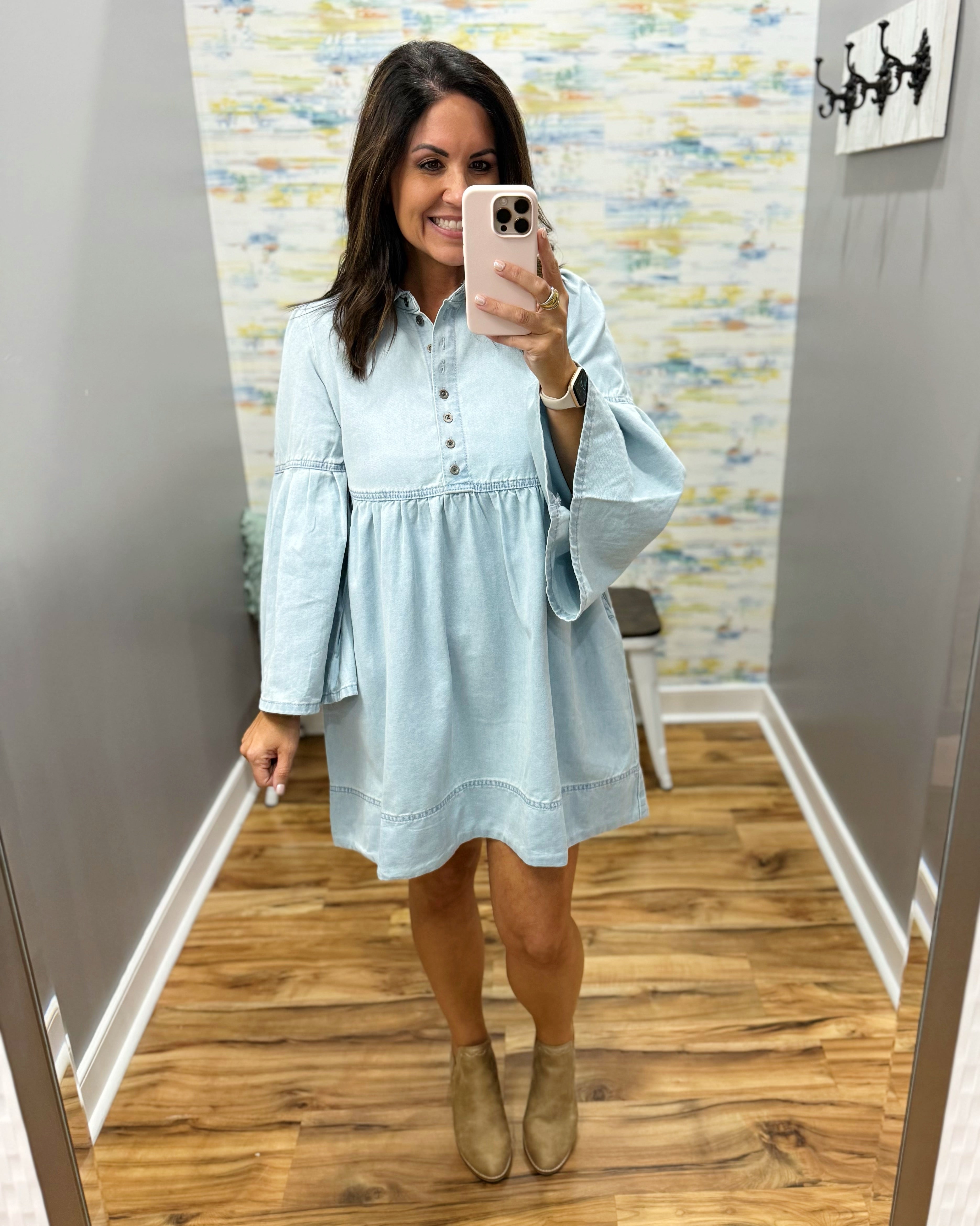 A woman takes a mirror selfie in a fitting room wearing a light blue long-sleeve button-up dress with a gathered waist and beige ankle boots.