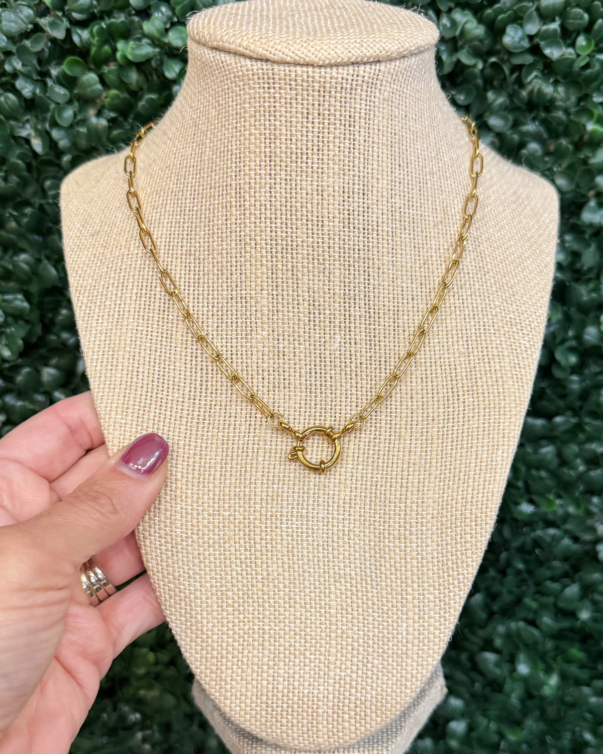 A gold chain necklace with a small interlocking ring clasp displayed on a beige fabric jewelry bust, with a hand holding the left side of the bust against a green leafy background.