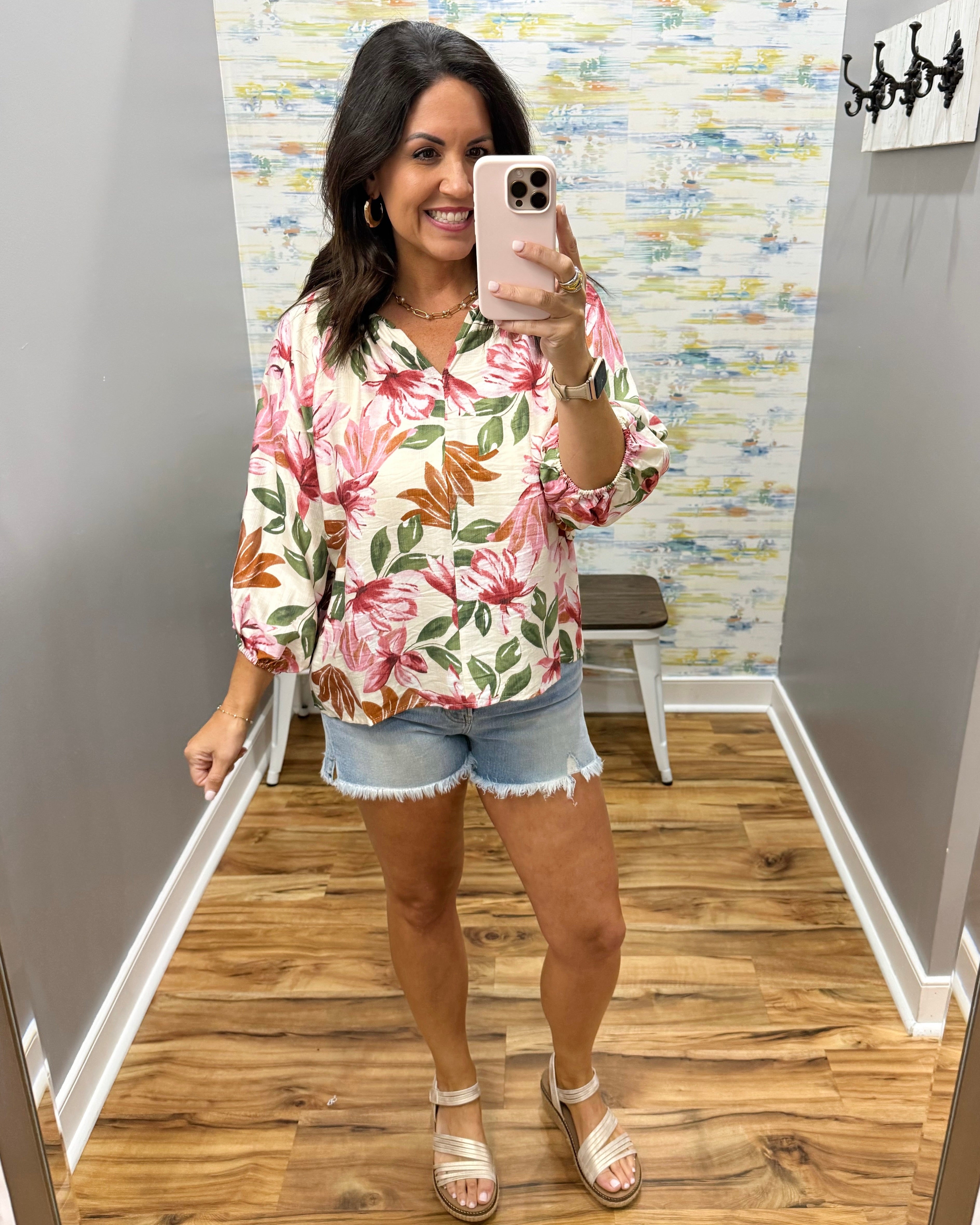Woman taking a mirror selfie wearing a floral blouse and denim shorts in a room with a patterned wall.