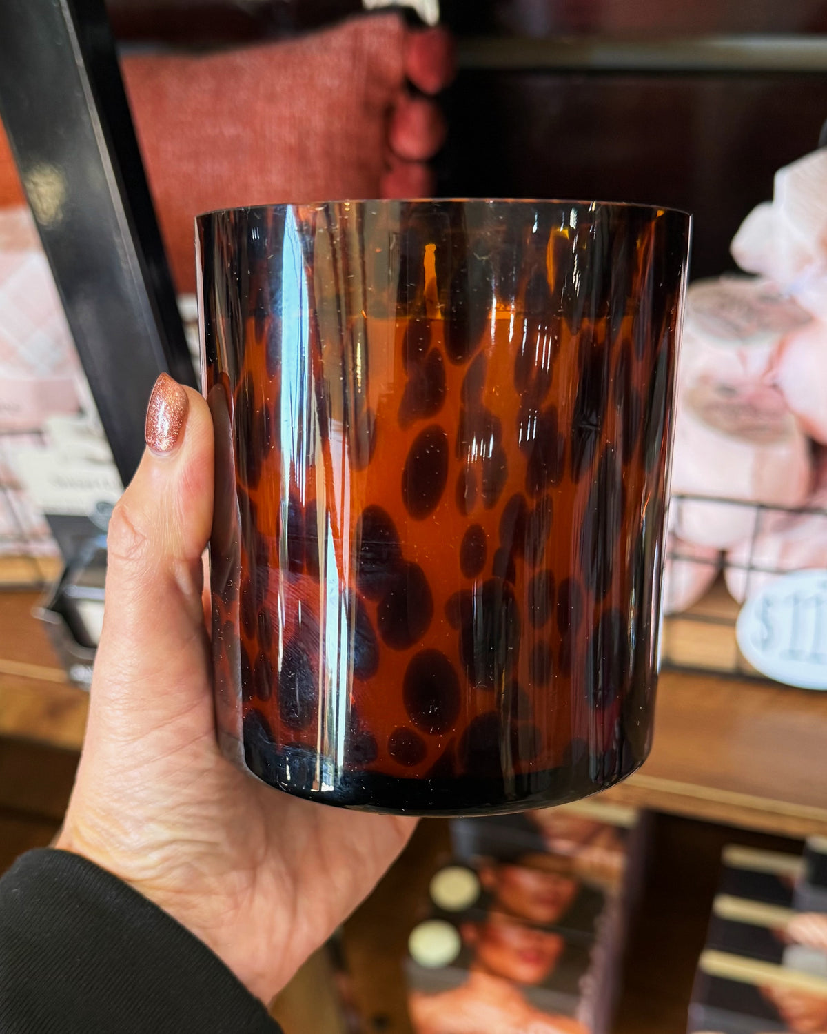 A hand holding a tall, cylindrical glass candle jar in sweet grace scent with a glossy amber-brown and black leopard print pattern, set against a blurred store background.