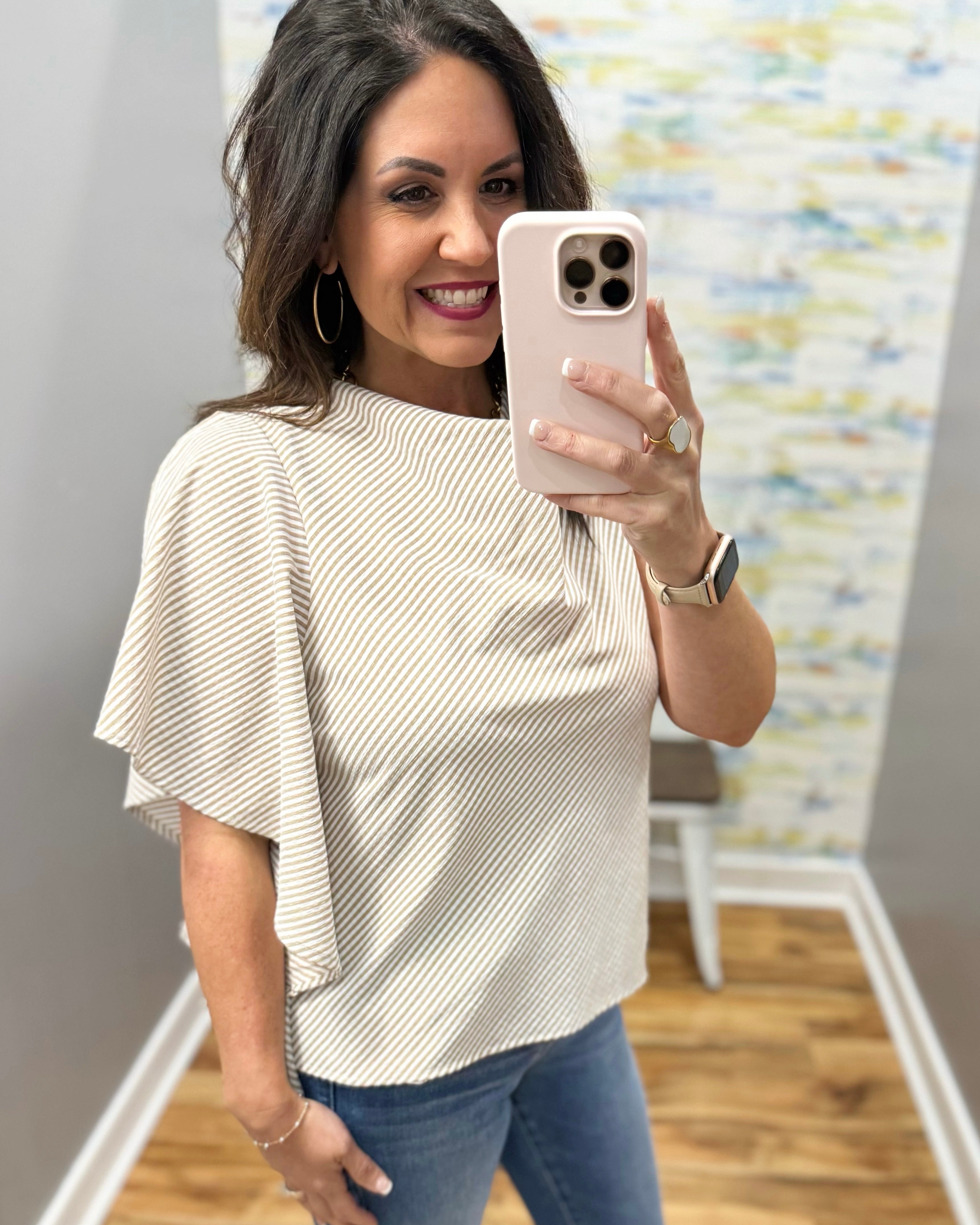 Woman taking a mirror selfie wearing a beige top and blue jeans in a room with a colorful abstract painting on the wall.