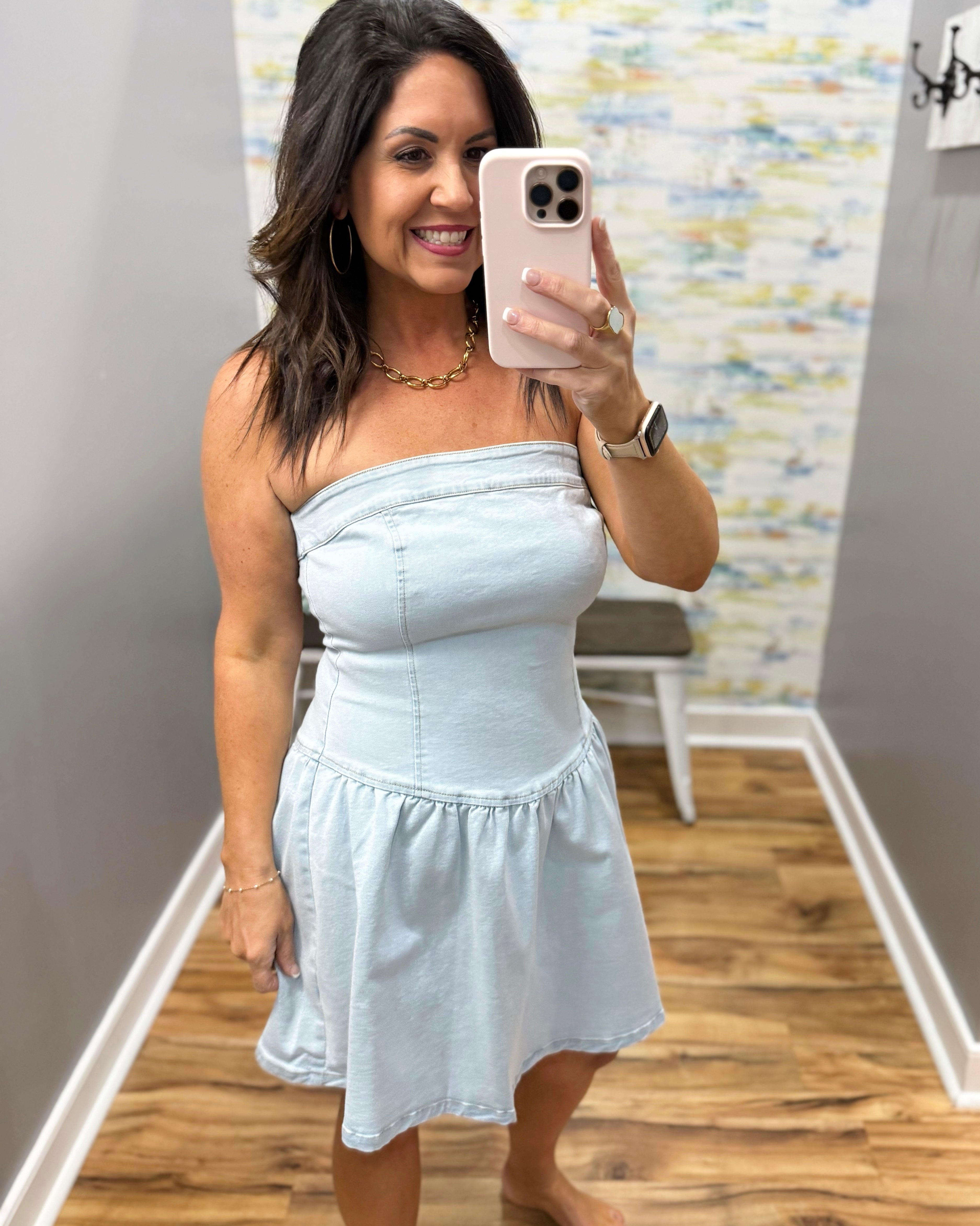 Woman taking a mirror selfie wearing a light blue strapless dress in a room with patterned wallpaper.