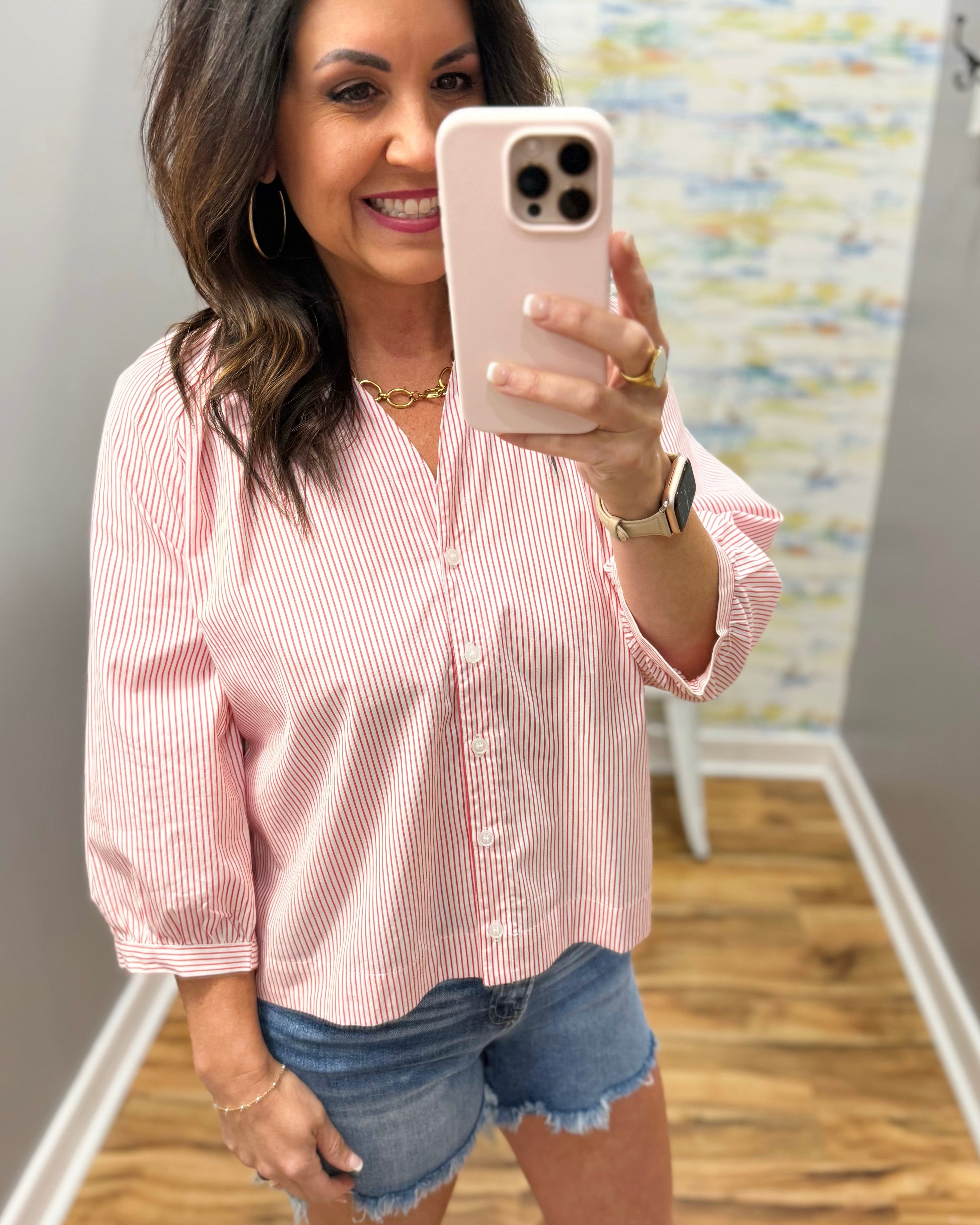 Woman taking a selfie in a pink striped shirt and denim shorts indoors.