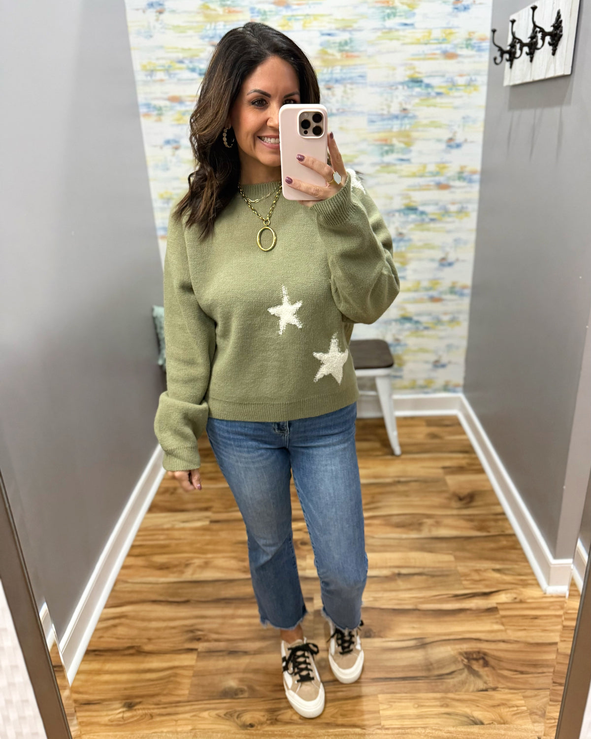 A woman takes a mirror selfie wearing an olive green knit sweater with white star patterns, blue jeans, and white sneakers with black laces in a home hallway.