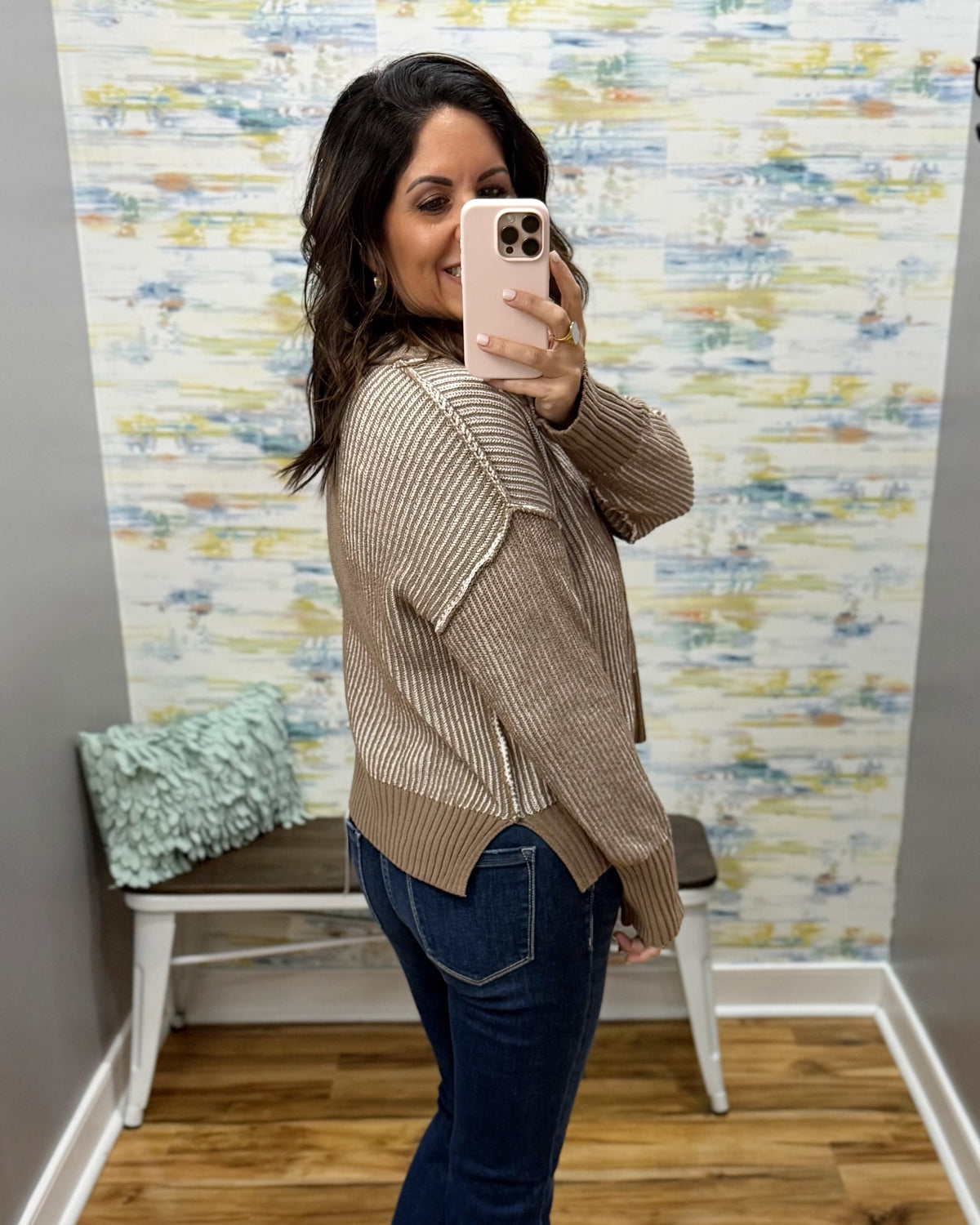 A woman takes a mirror selfie wearing a beige ribbed knit cardigan sweater and dark blue jeans in a room with a patterned wallpaper background.