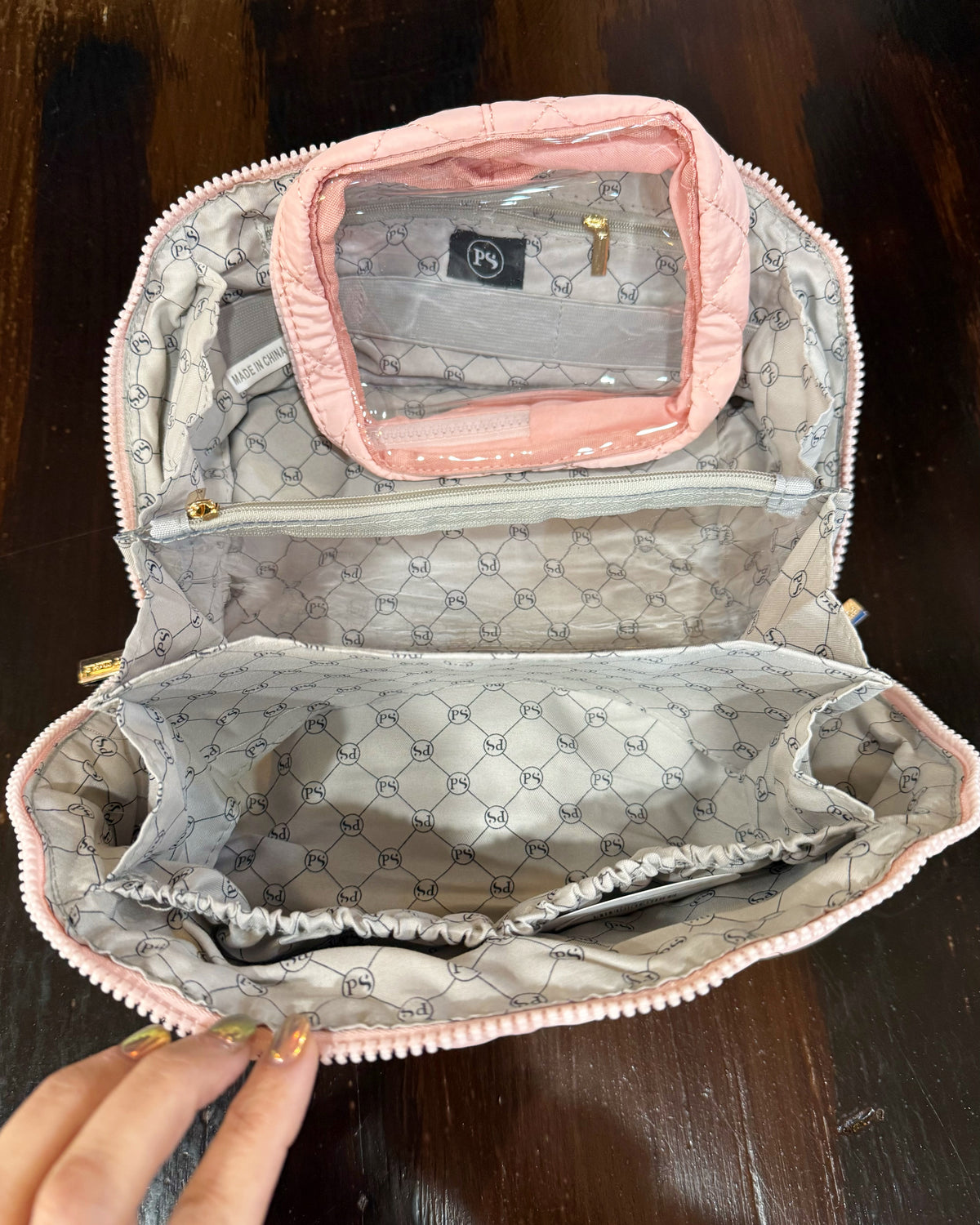 An open quilted cosmetic bag with a pink exterior and a gray and white geometric monogram pattern lining, displayed on a dark wooden surface.