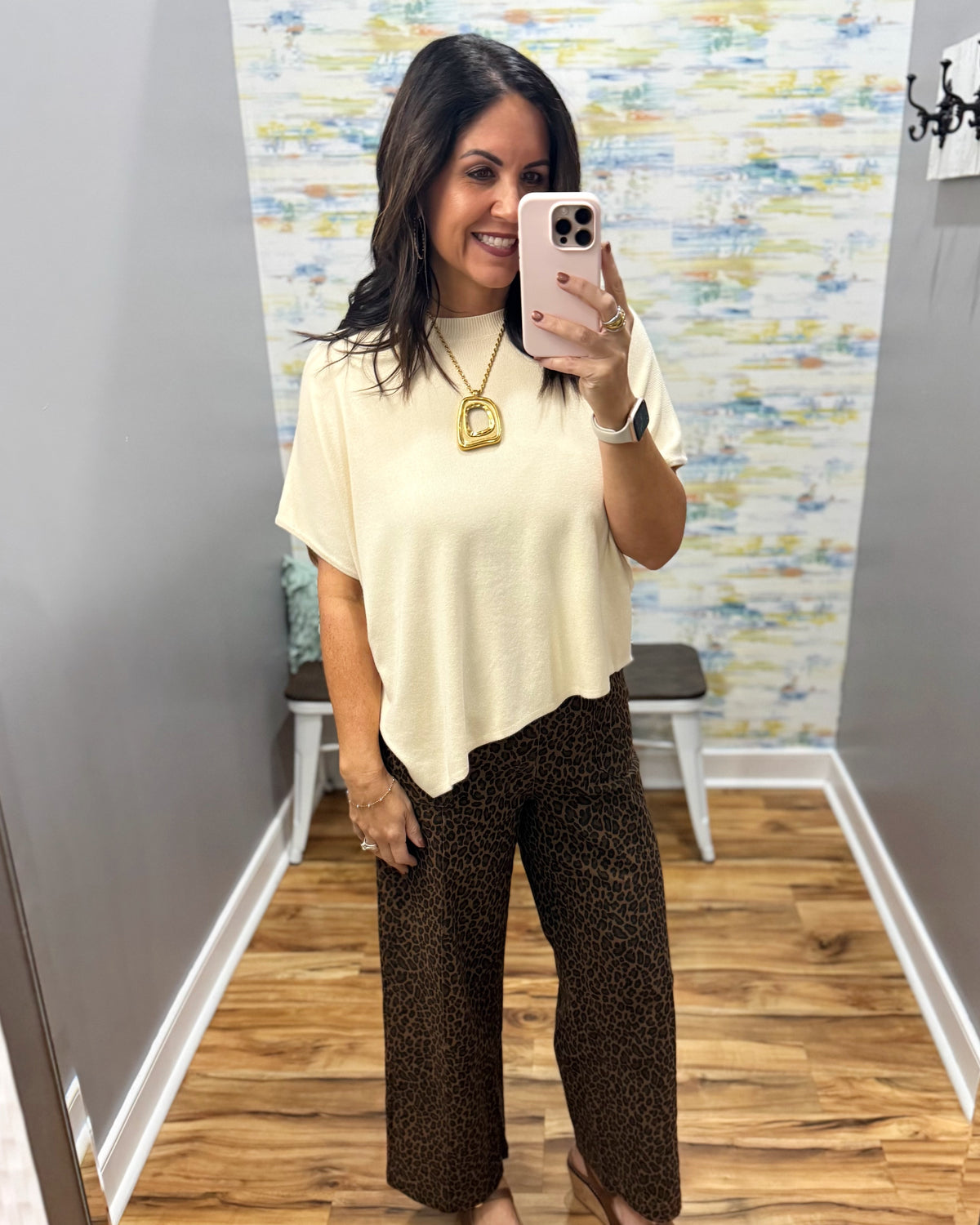 A woman takes a mirror selfie in a fitting room wearing a cream short-sleeve top and brown leopard-print pants. She is wearing a gold pendant necklace and holding a white phone.
