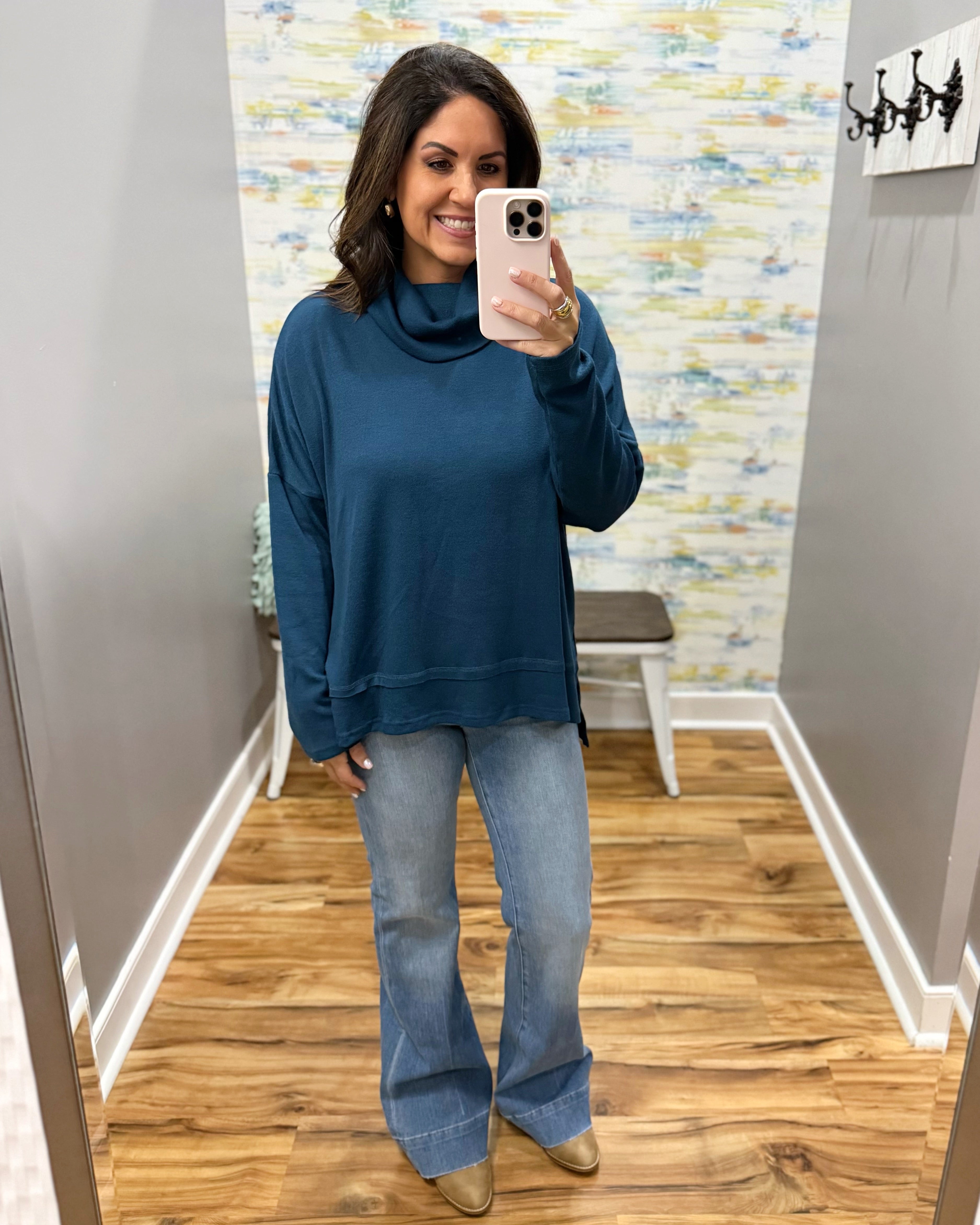 A woman takes a mirror selfie in a fitting room wearing a dark teal blue turtleneck sweatshirt and light-wash blue flare jeans with tan pointed-toe flats.