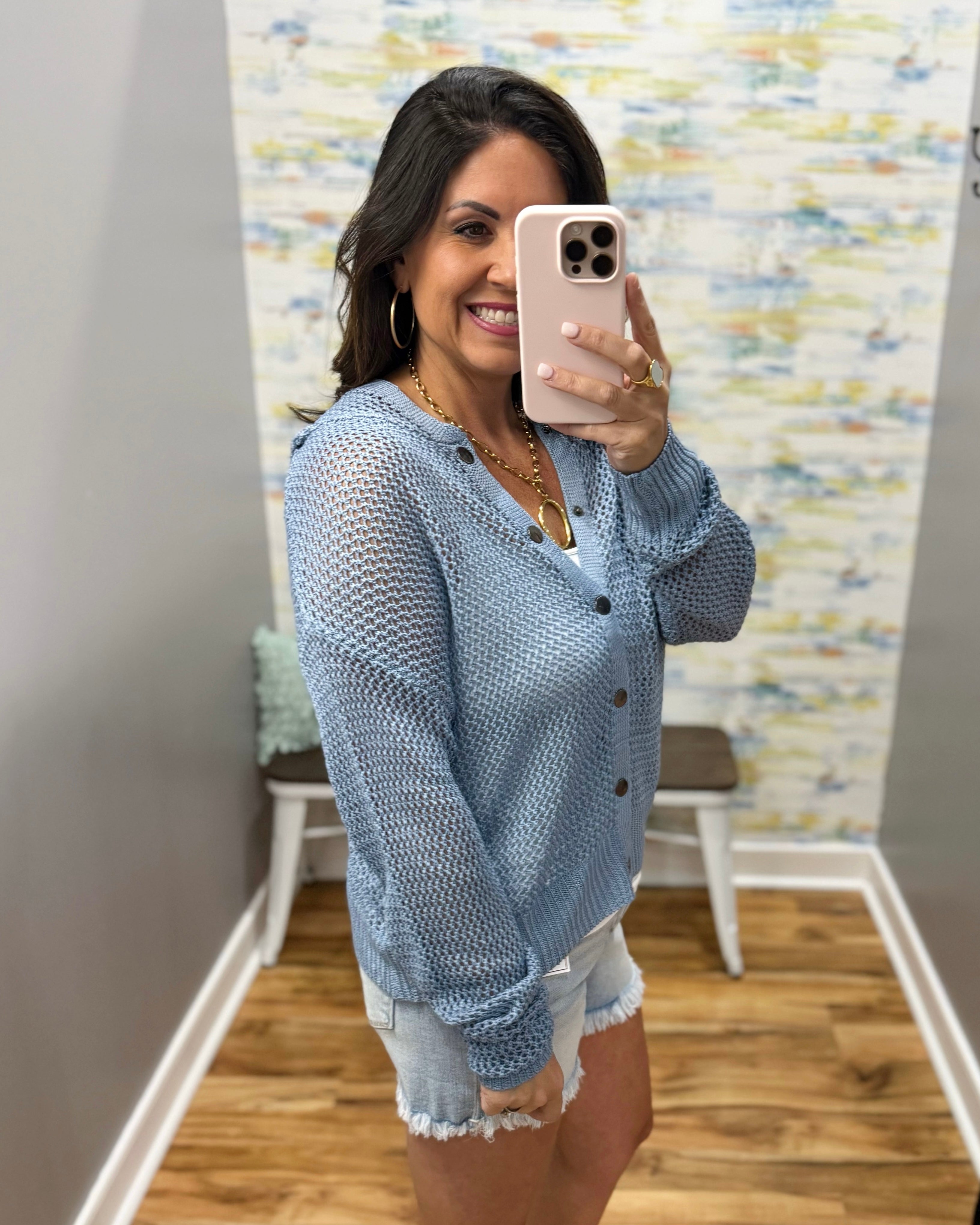 Woman taking a mirror selfie wearing a light blue cardigan and white shorts in a room with a colorful wall.