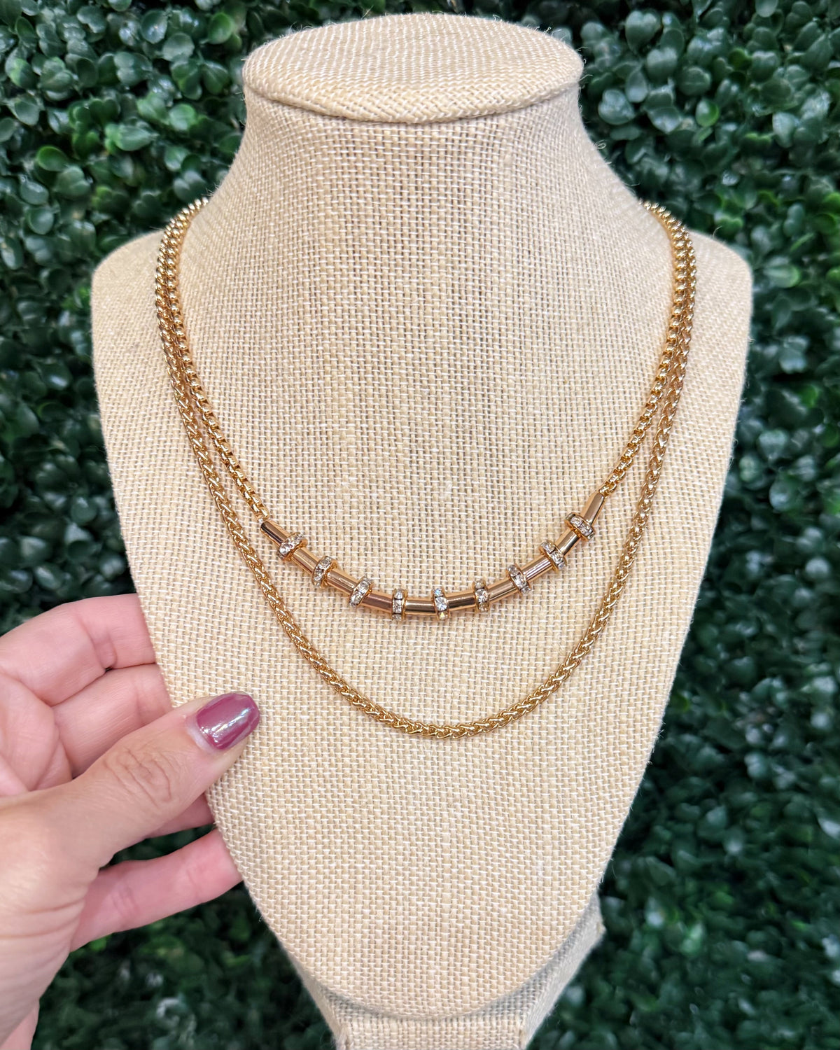 A gold-toned double-strand necklace displayed on a beige fabric jewelry bust, with a hand holding the left side of the bust. The necklace features two chains with a central row of small silver-toned square links.