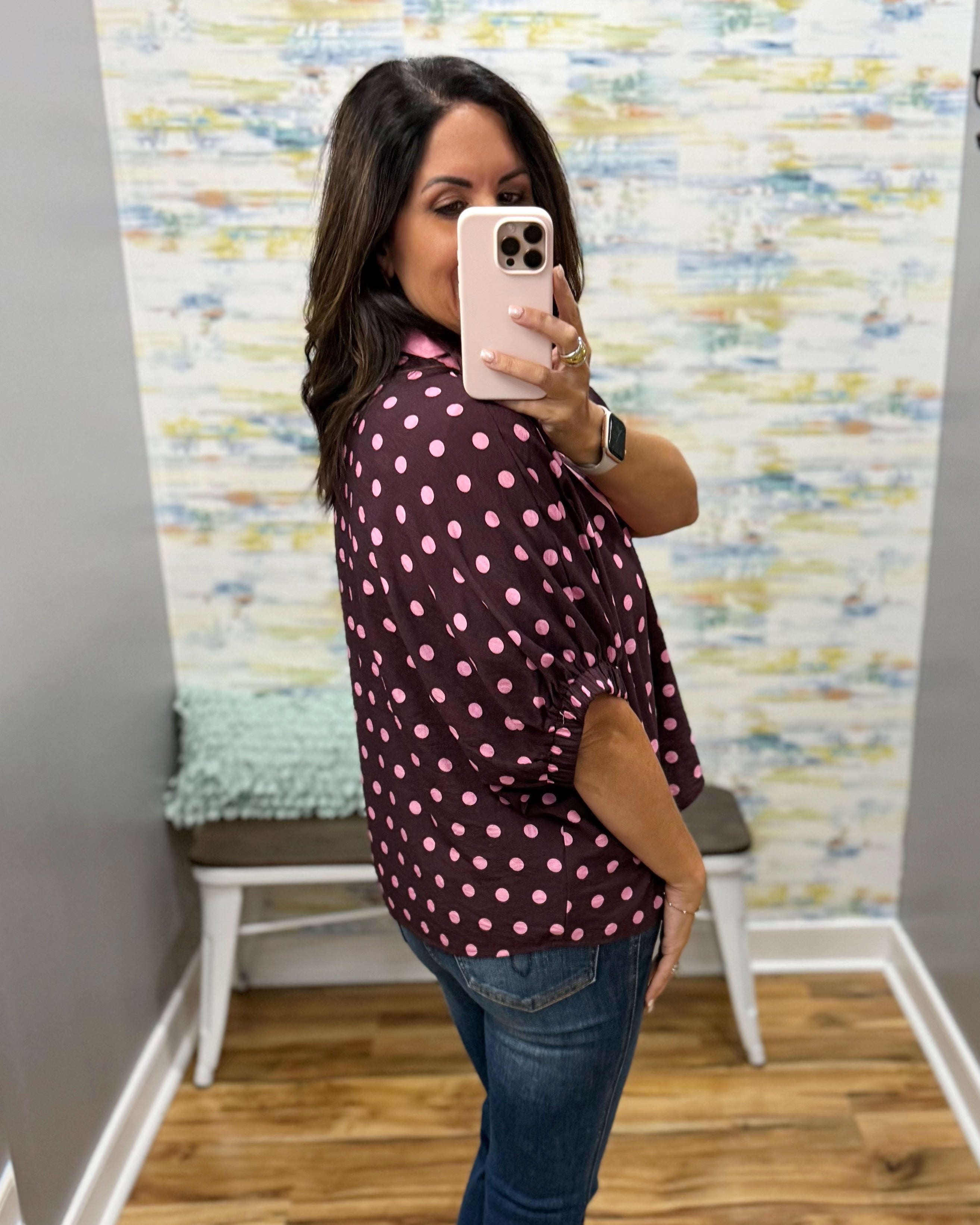 A woman takes a mirror selfie wearing a dark burgundy button-up blouse with pink polka dots and blue jeans, standing in a room with a patterned wall and wooden floor.