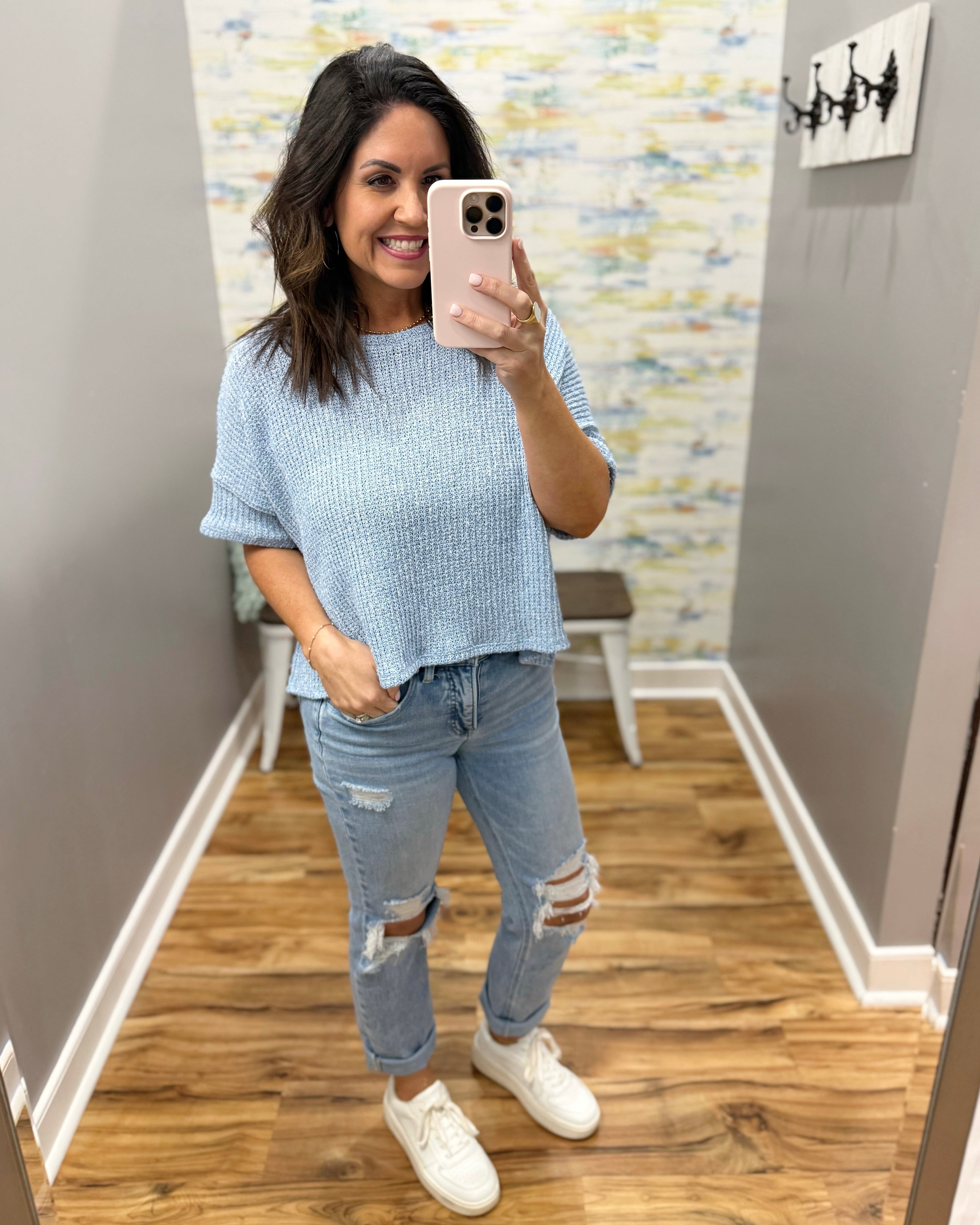Woman taking a mirror selfie wearing a light blue sweater and ripped jeans in a room with a patterned wall.