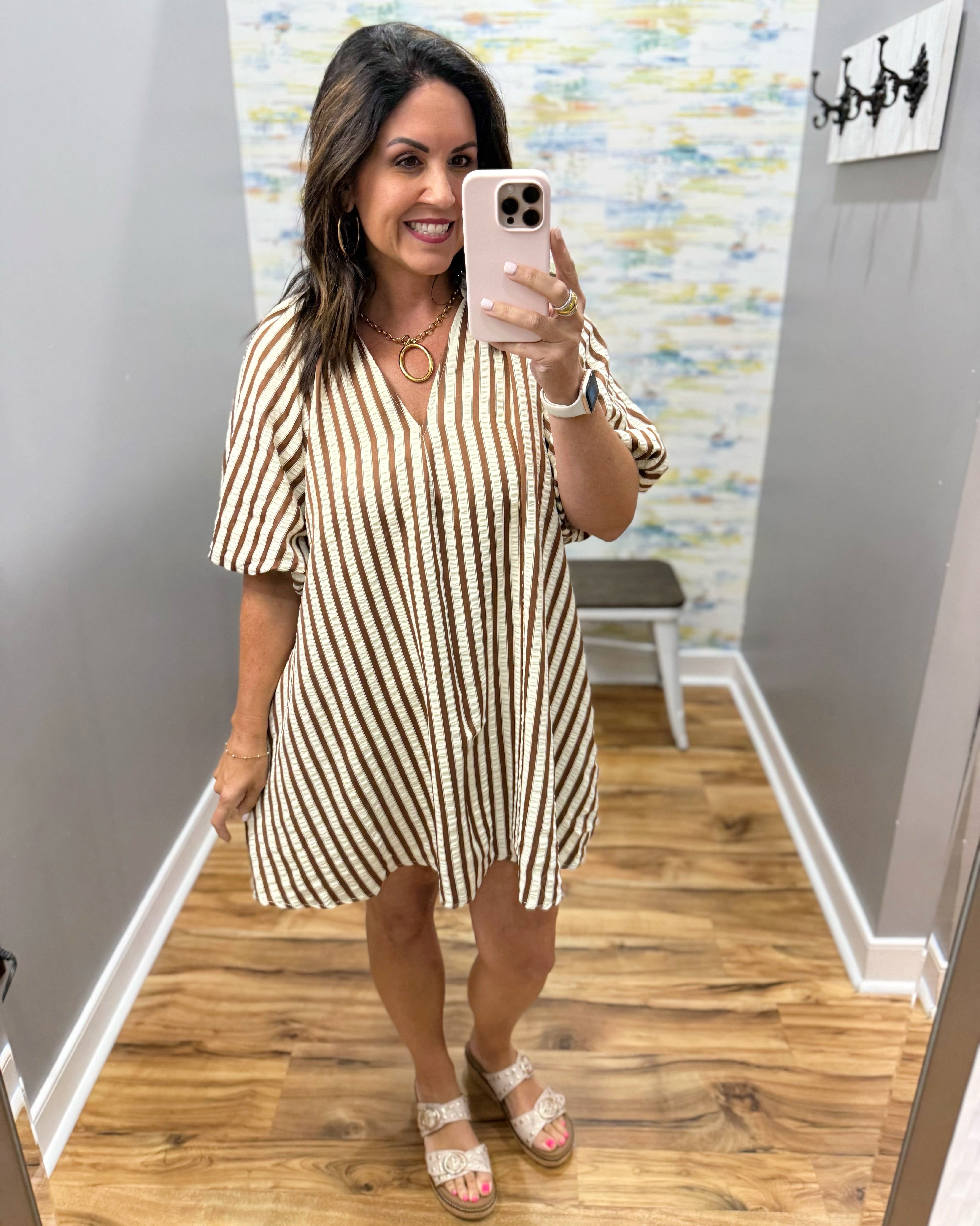 A woman takes a mirror selfie in a fitting room wearing a short-sleeve V-neck mini dress with vertical brown and cream stripes, paired with white lace-up sandals.