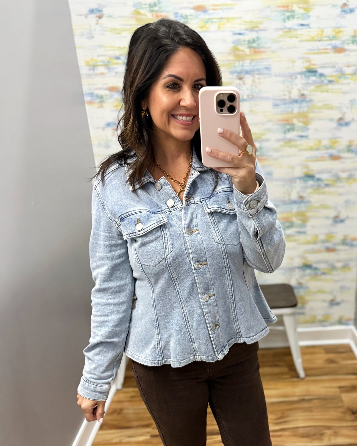 A woman takes a mirror selfie wearing a light-wash blue denim cropped jacket and dark brown pants in an indoor setting with a patterned wall and wood floor.