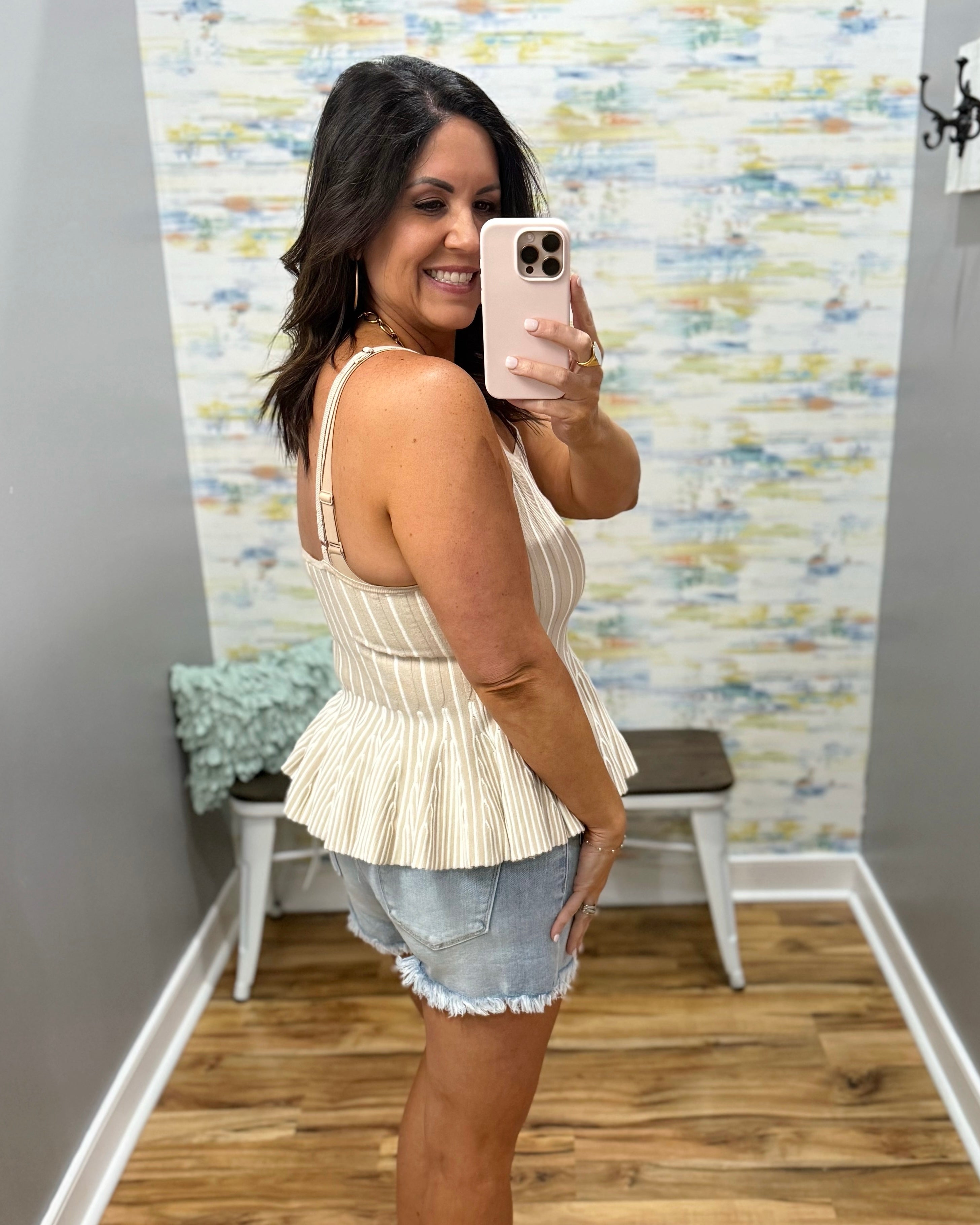 Woman taking a mirror selfie wearing a beige peplum top and denim shorts in a room with a textured wall.