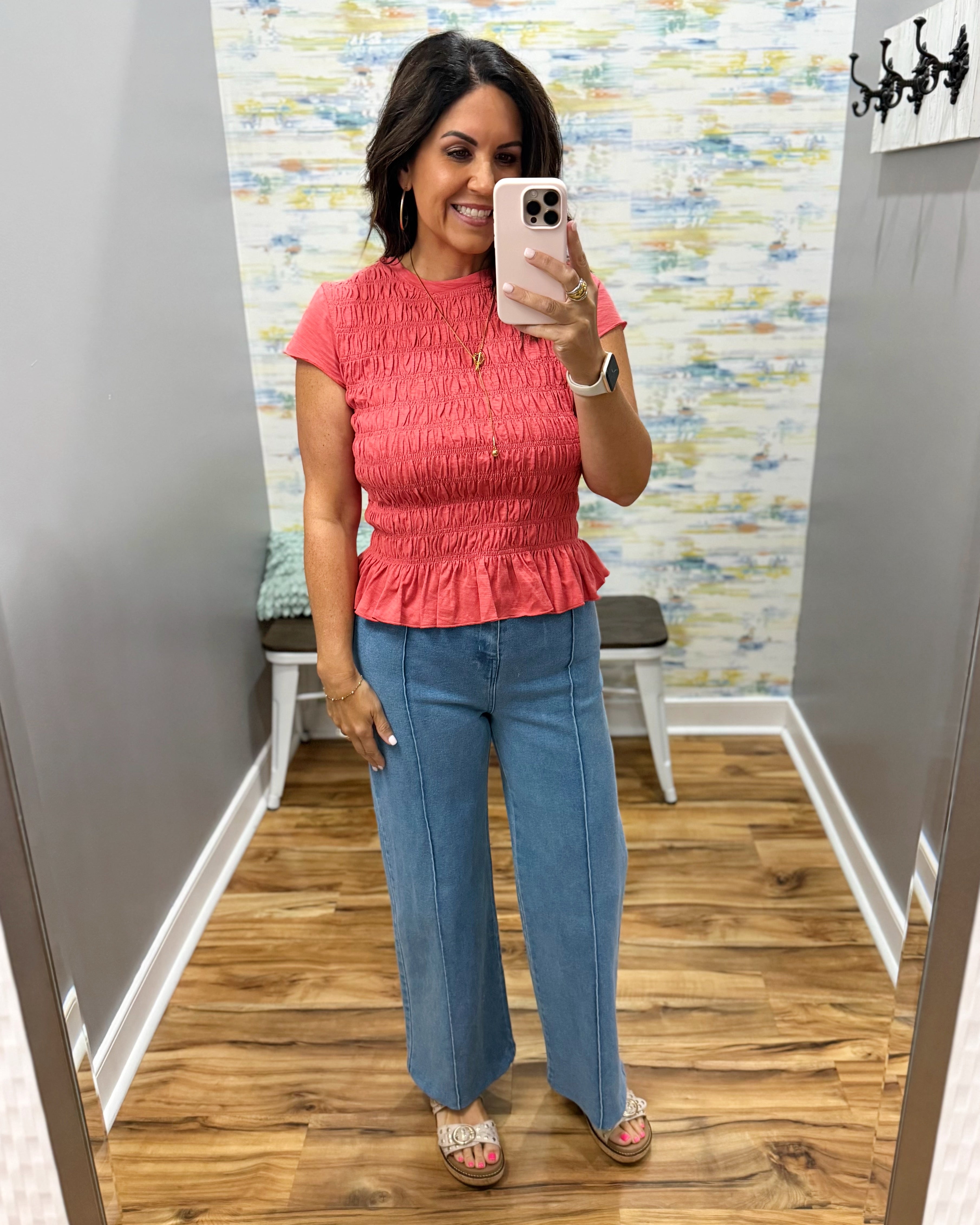 A woman takes a mirror selfie wearing a coral pink short-sleeve smocked peplum top and blue jeans in a fitting room.