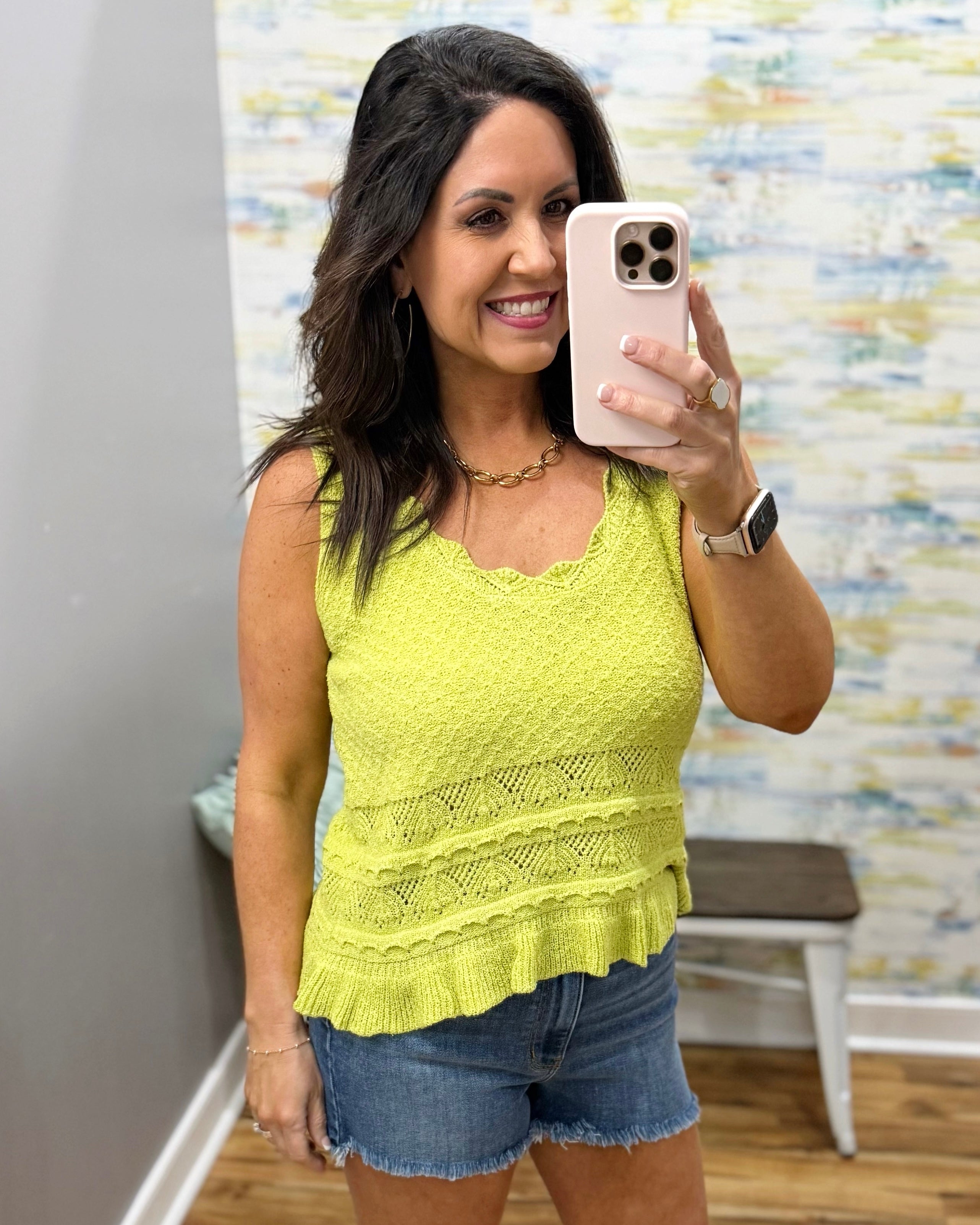 Woman taking a mirror selfie wearing a yellow top and denim shorts in a room with a patterned wall.