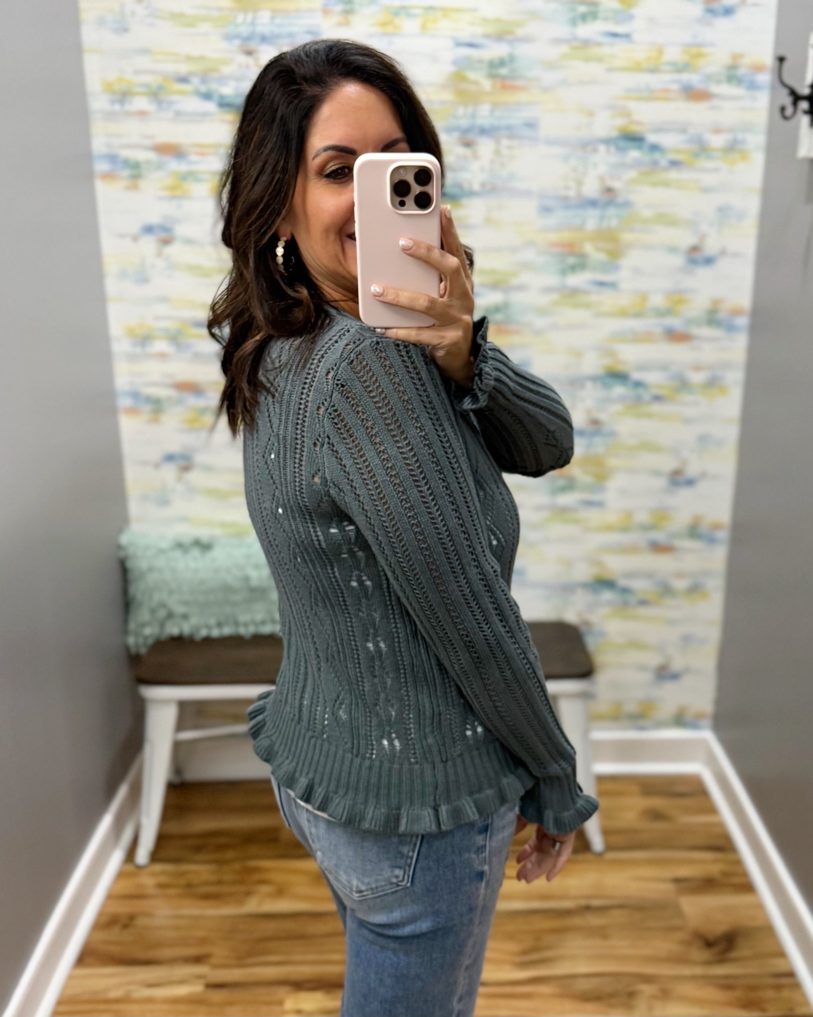 A woman takes a mirror selfie in a fitting room wearing a dark gray ribbed knit sweater with small white embroidered details, paired with blue jeans.