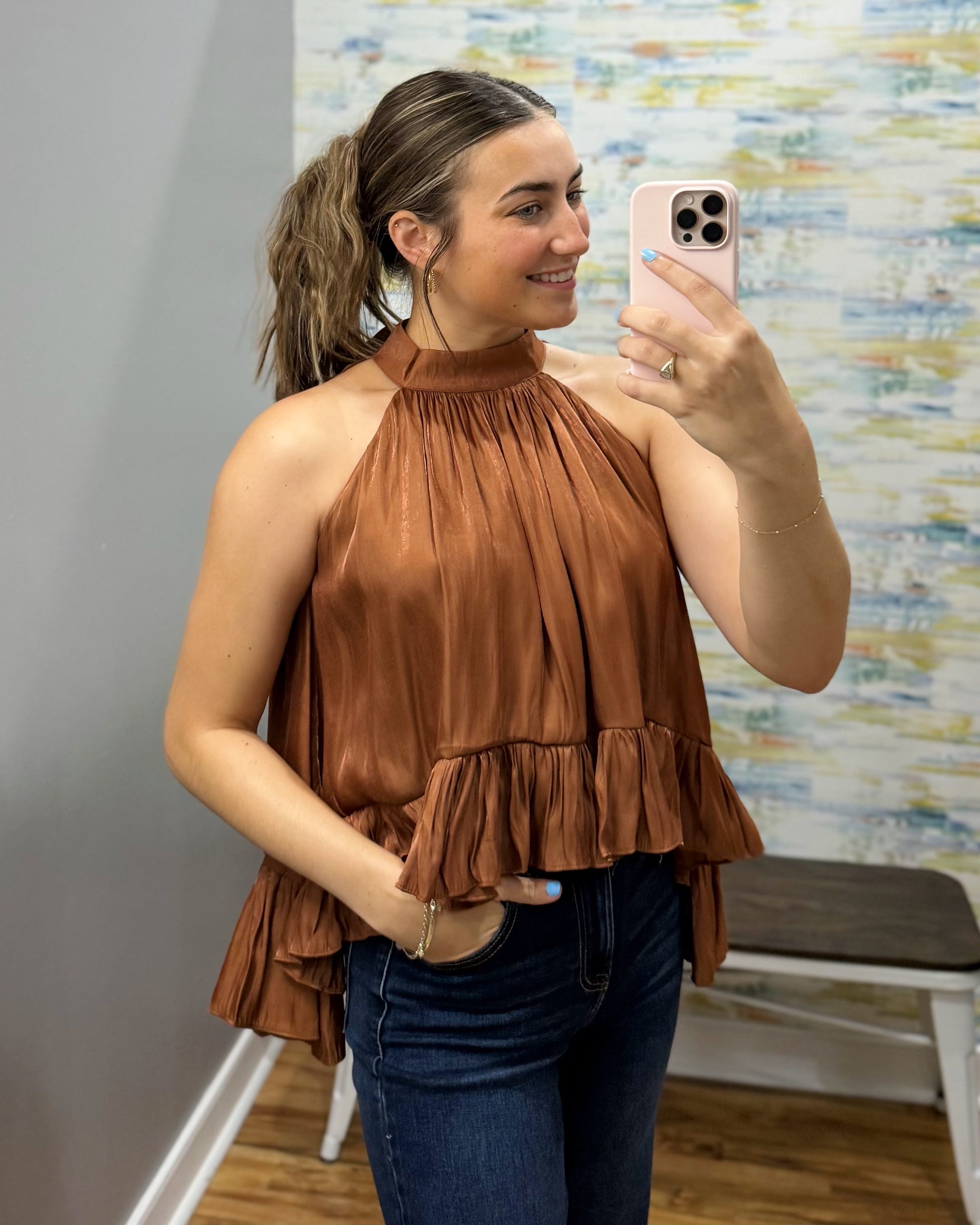A woman takes a mirror selfie wearing a brown halter-neck ruffled top and dark blue jeans in a fitting room.
