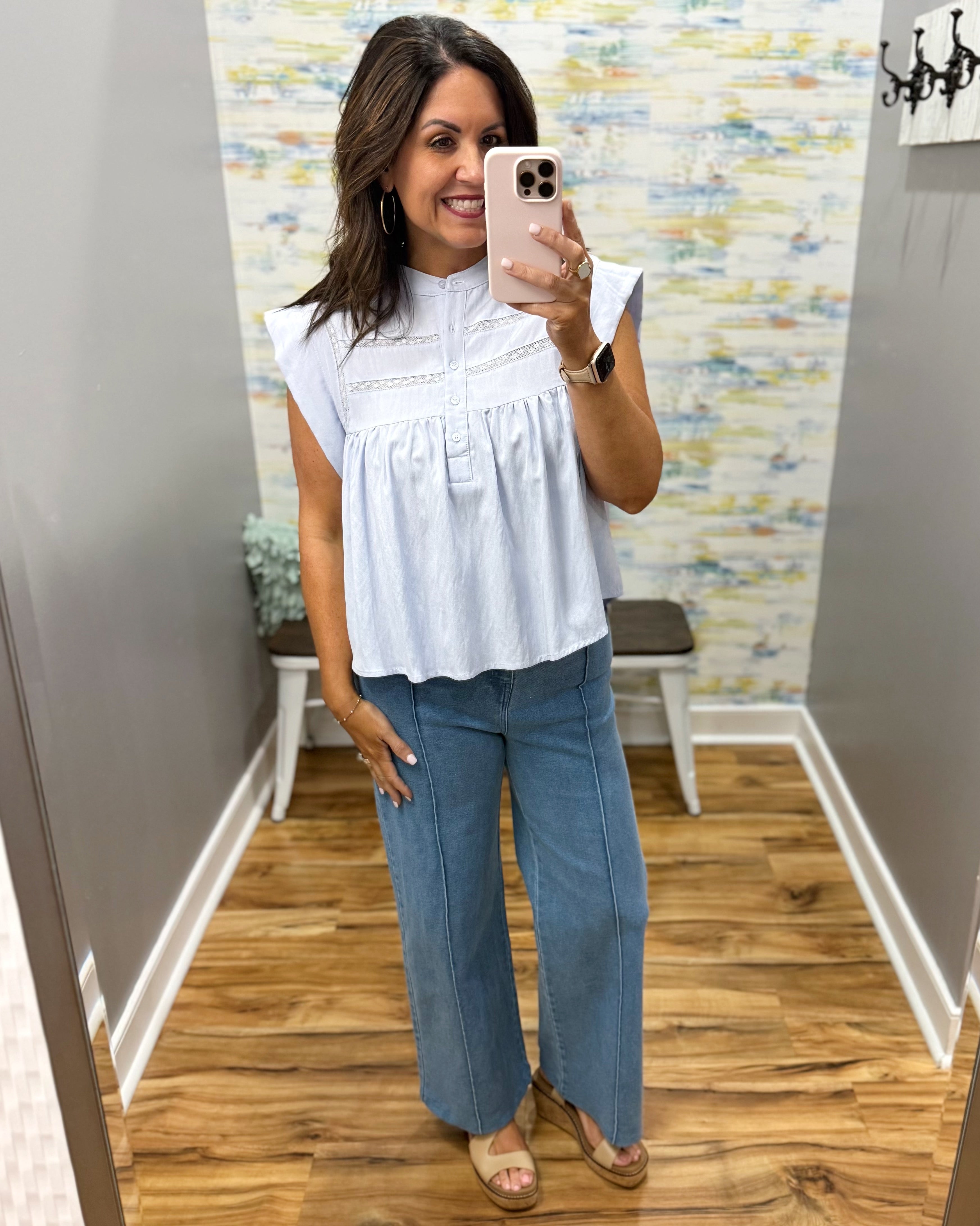 Woman taking a mirror selfie wearing a light blue top and blue jeans in a room with a patterned wall.