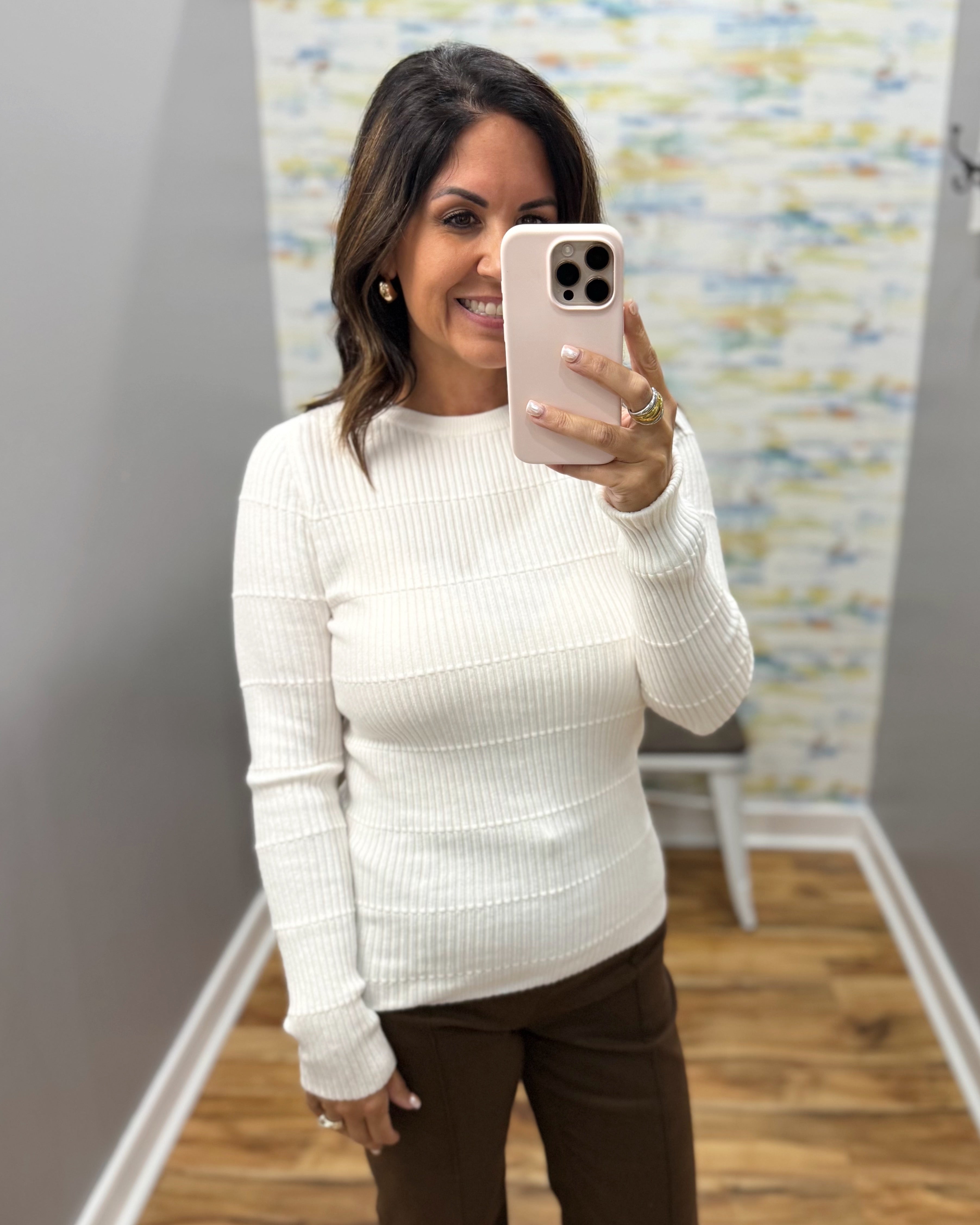 A woman taking a mirror selfie wearing a cream ribbed knit long-sleeve sweater and brown pants, holding a light pink smartphone.