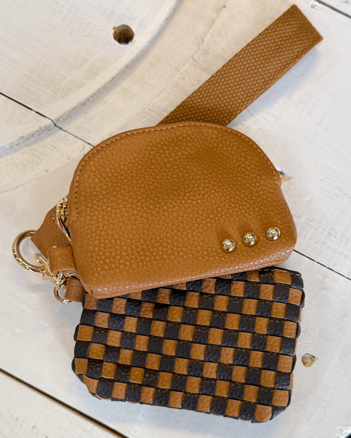 A small tan pebbled leather pouch with a wrist strap and gold-tone hardware, layered over a woven brown-and-black check pouch, on a white wooden surface.