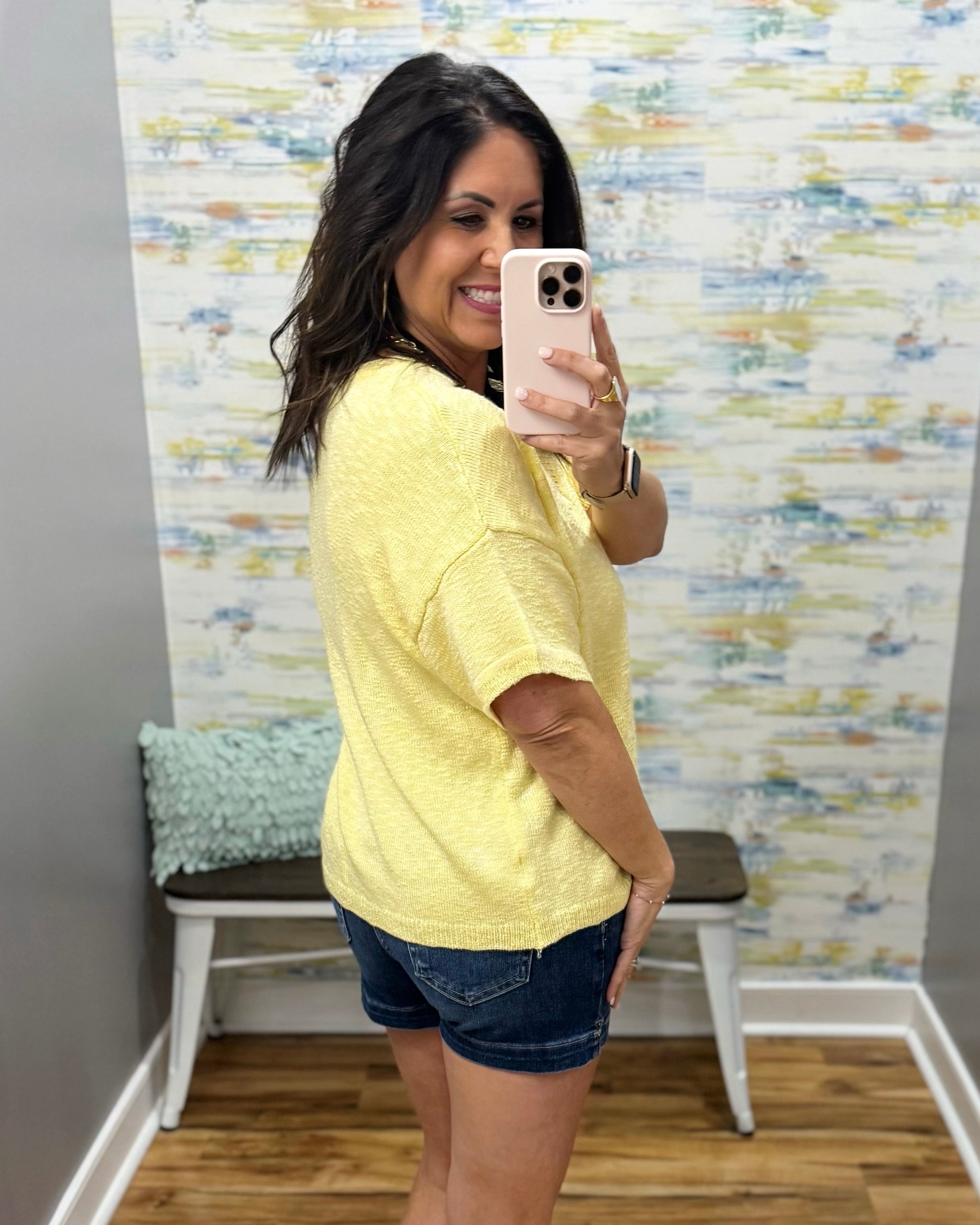 Woman taking a mirror selfie wearing a yellow top and denim shorts in a room with colorful wall paper.