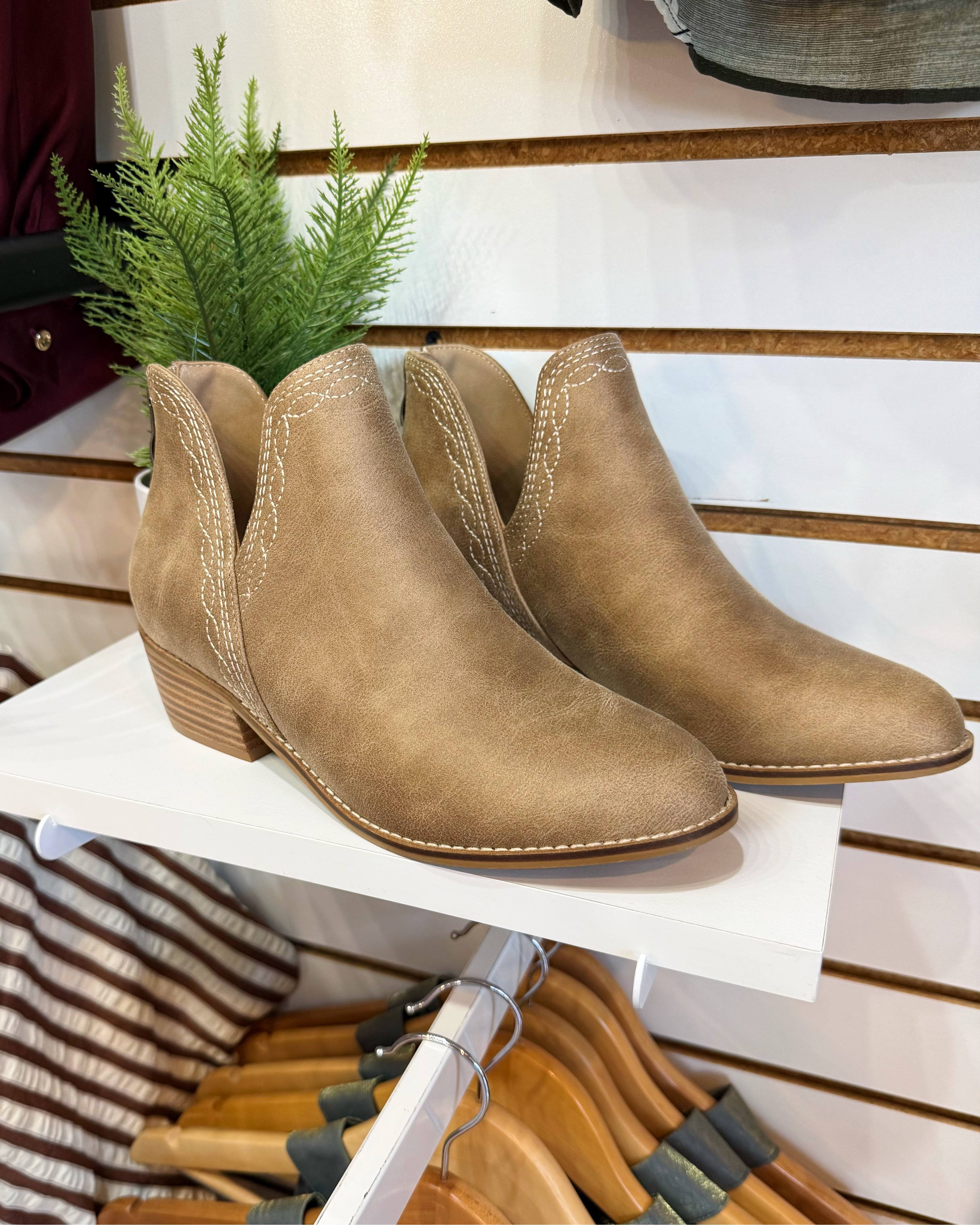 A pair of taupe-brown suede ankle boots with stacked block heels, displayed on a white shelf in a retail setting.