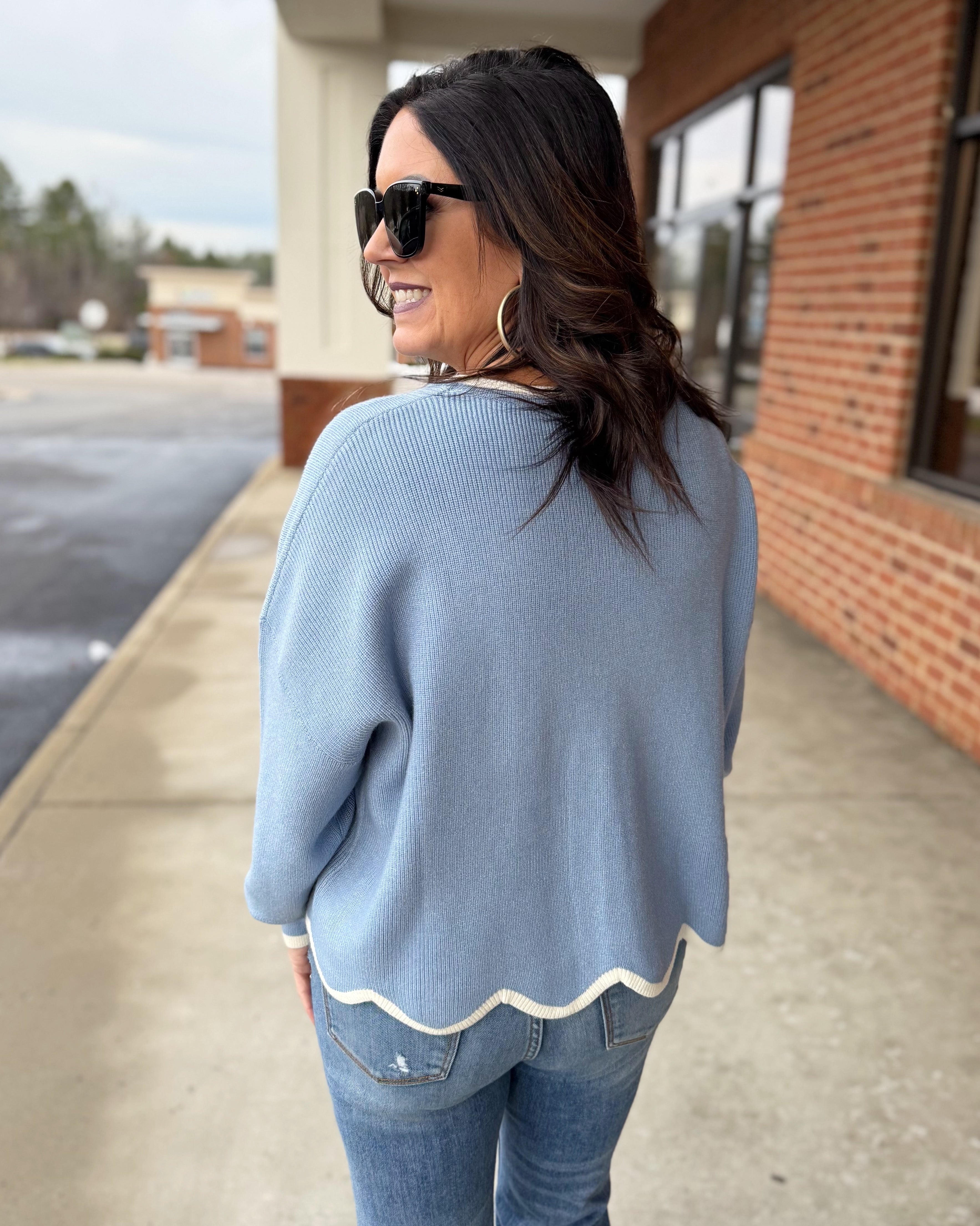 Woman wearing a light blue sweater and sunglasses, standing on a sidewalk.