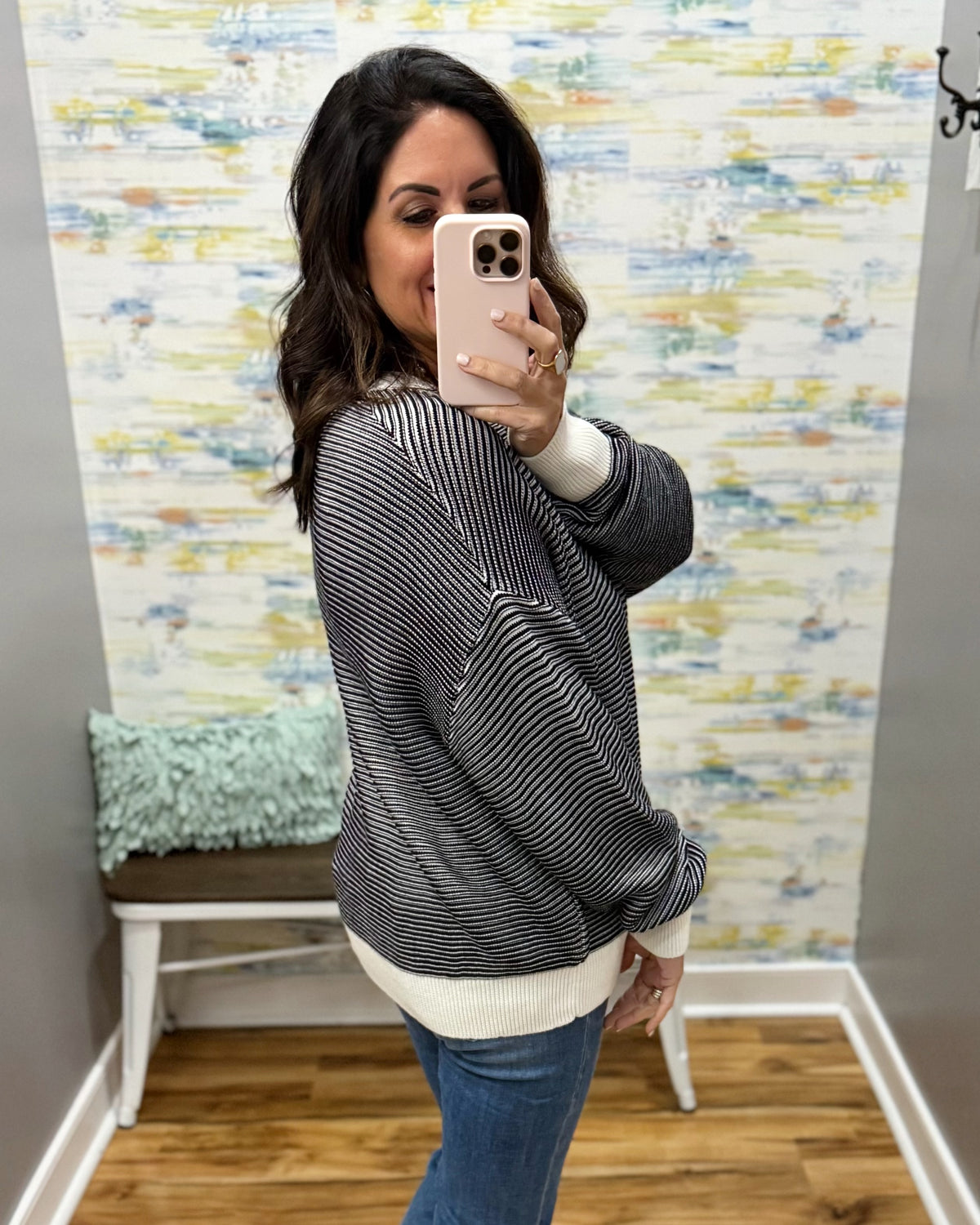 A woman takes a mirror selfie wearing a black and white striped knit sweater with a cream ribbed hem and cuffs, paired with blue jeans. The background features a colorful abstract wallpaper, a small bench with a mint green pillow, and a wood floor.