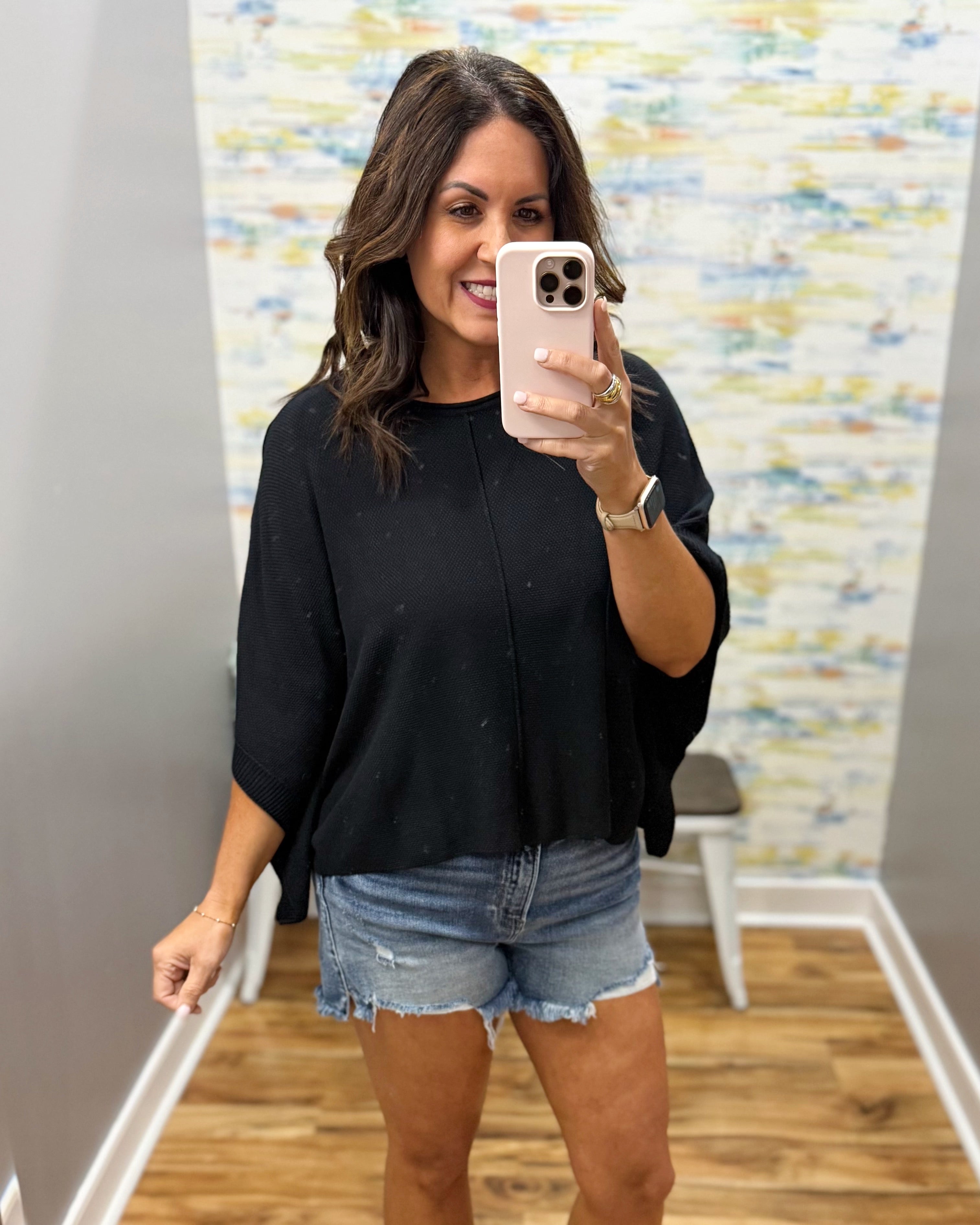 Woman taking a mirror selfie wearing a black top and denim shorts in a room with a colorful wall.