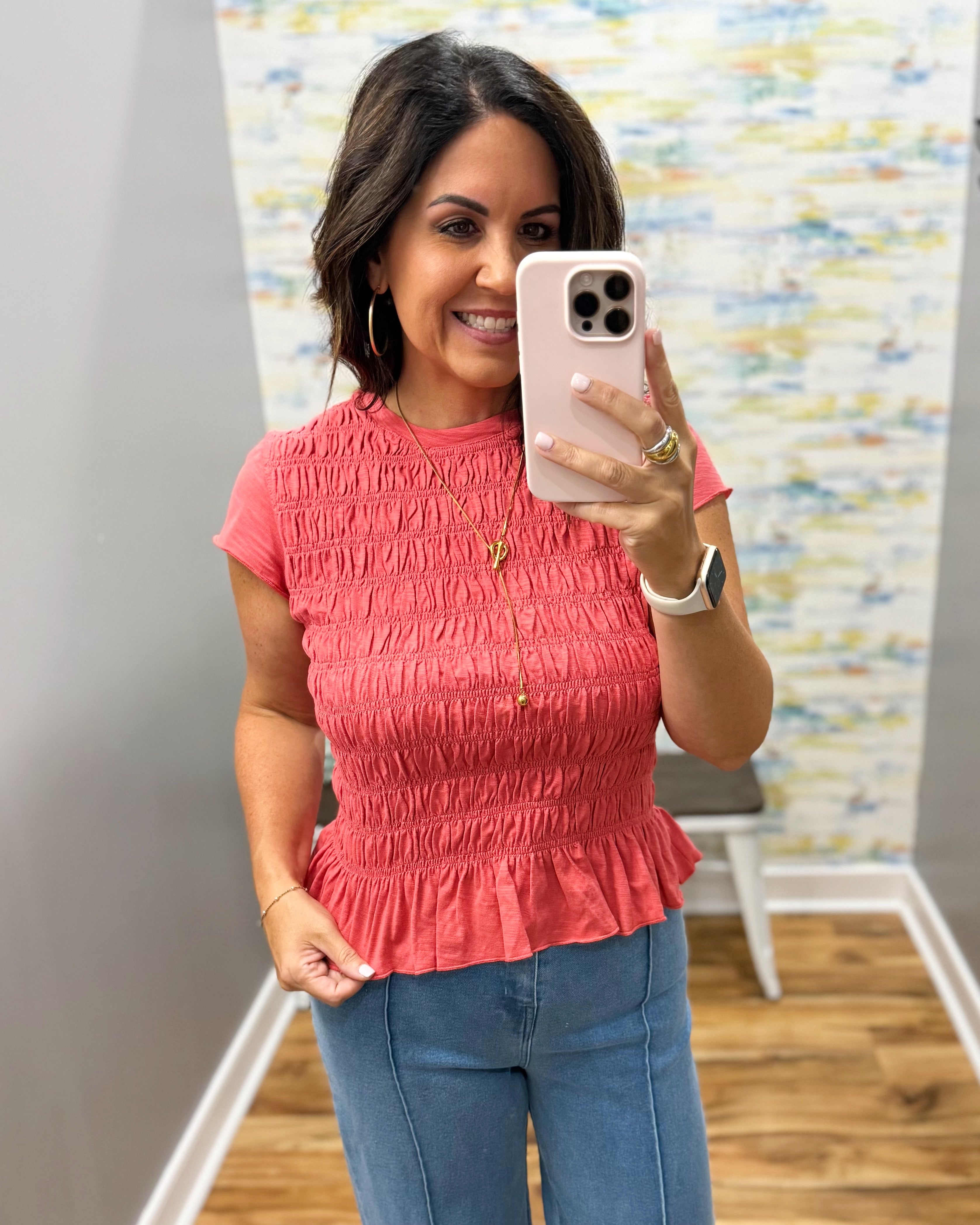 A woman takes a mirror selfie wearing a coral pink short-sleeve smocked peplum top and blue jeans in a fitting room.