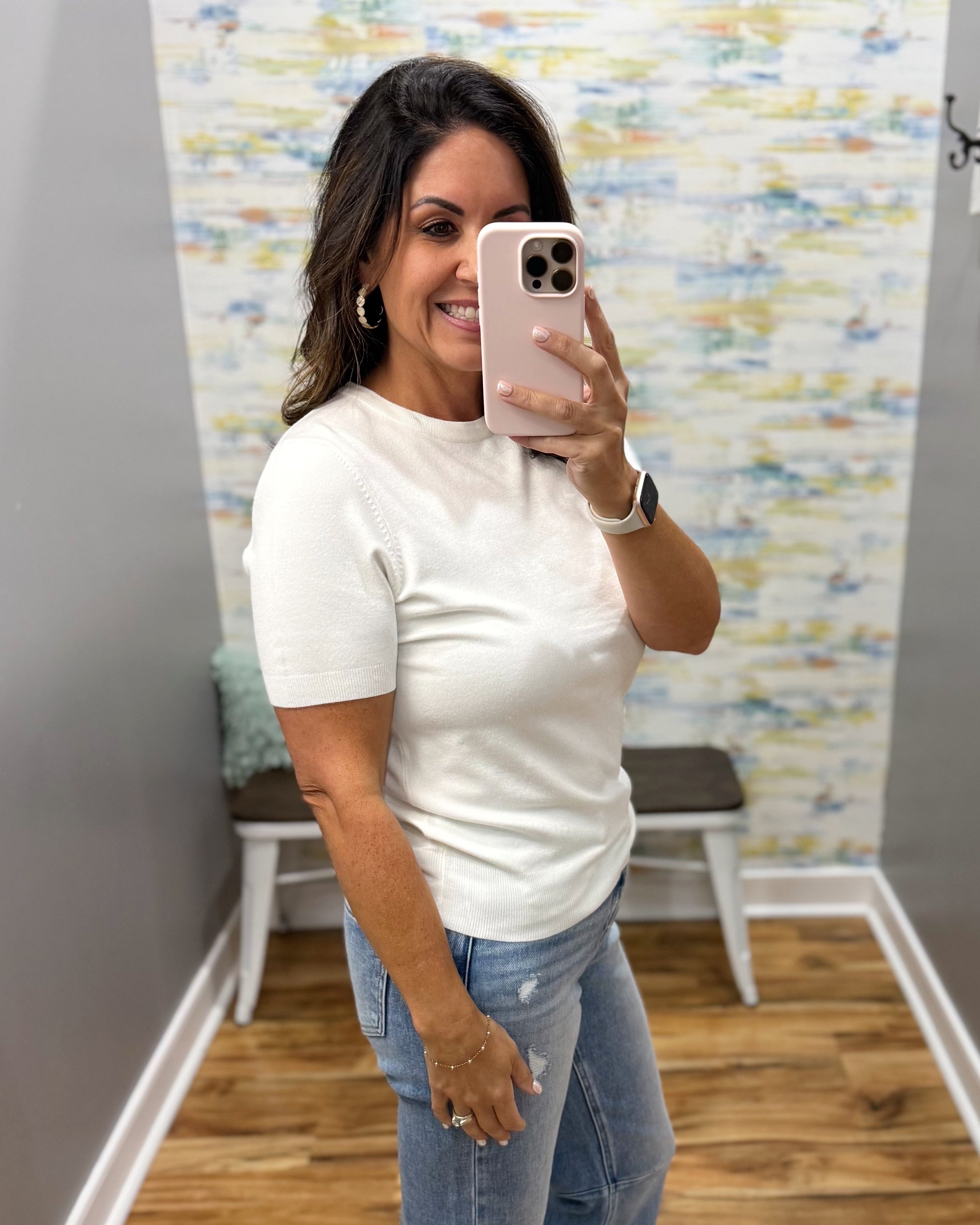 A woman takes a mirror selfie wearing a fitted white short-sleeve knit top and light-wash blue jeans in a fitting room with a patterned wall and wood floor.