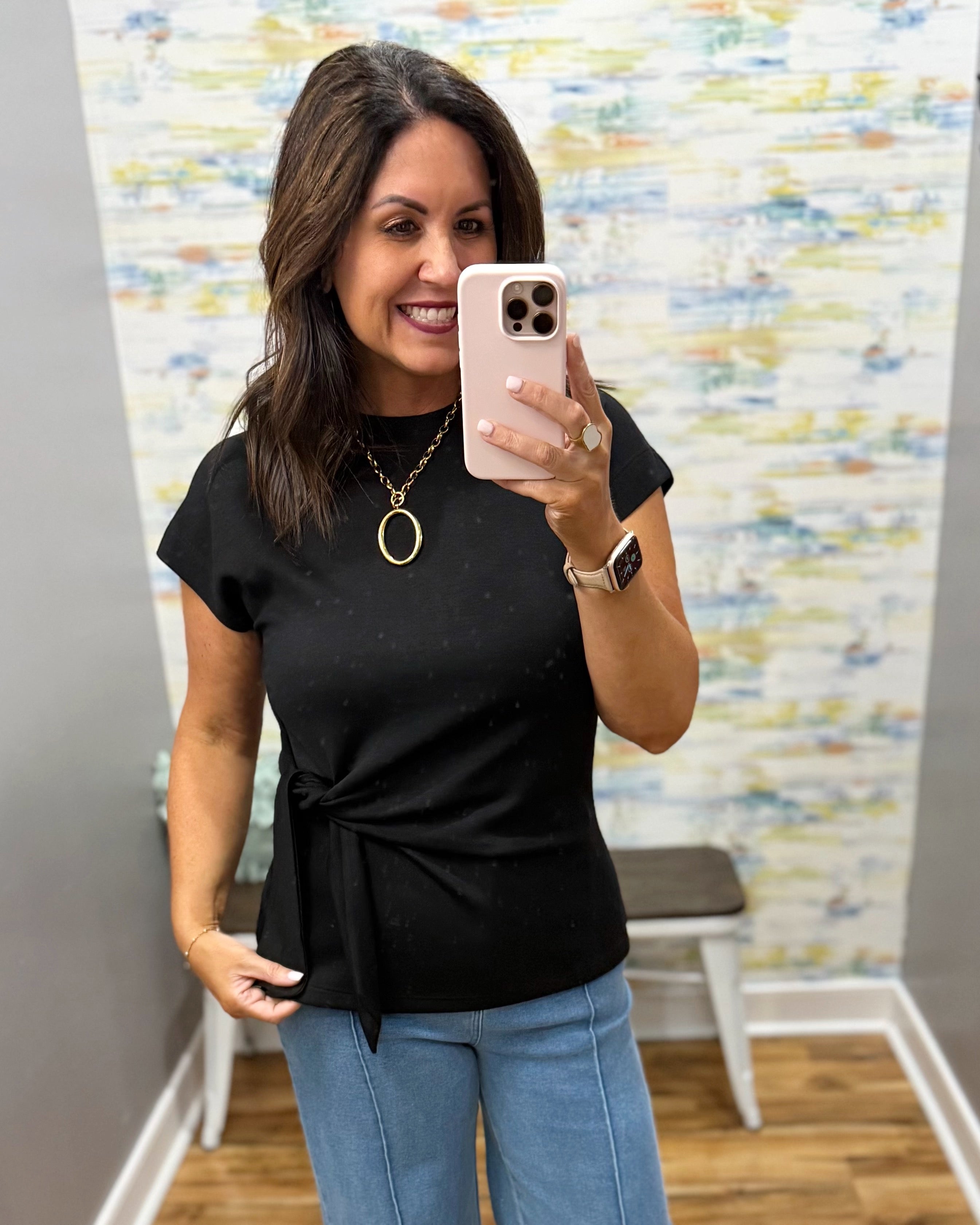 Woman taking a mirror selfie wearing a black top and blue jeans in a room with colorful wall art.
