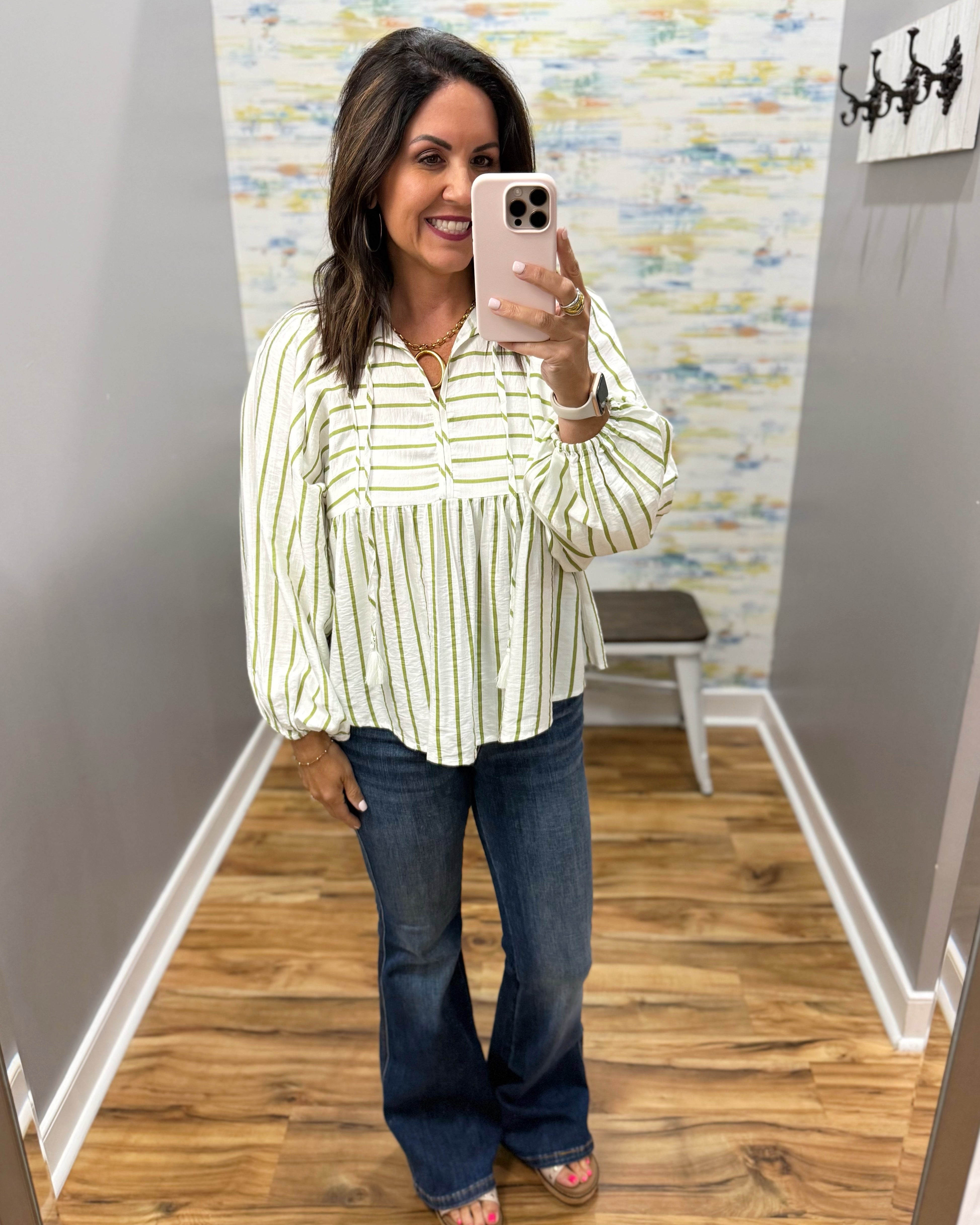 A woman takes a mirror selfie in a fitting room wearing a cream and green vertically striped long-sleeve blouse with a peplum waist, paired with dark blue flare jeans.