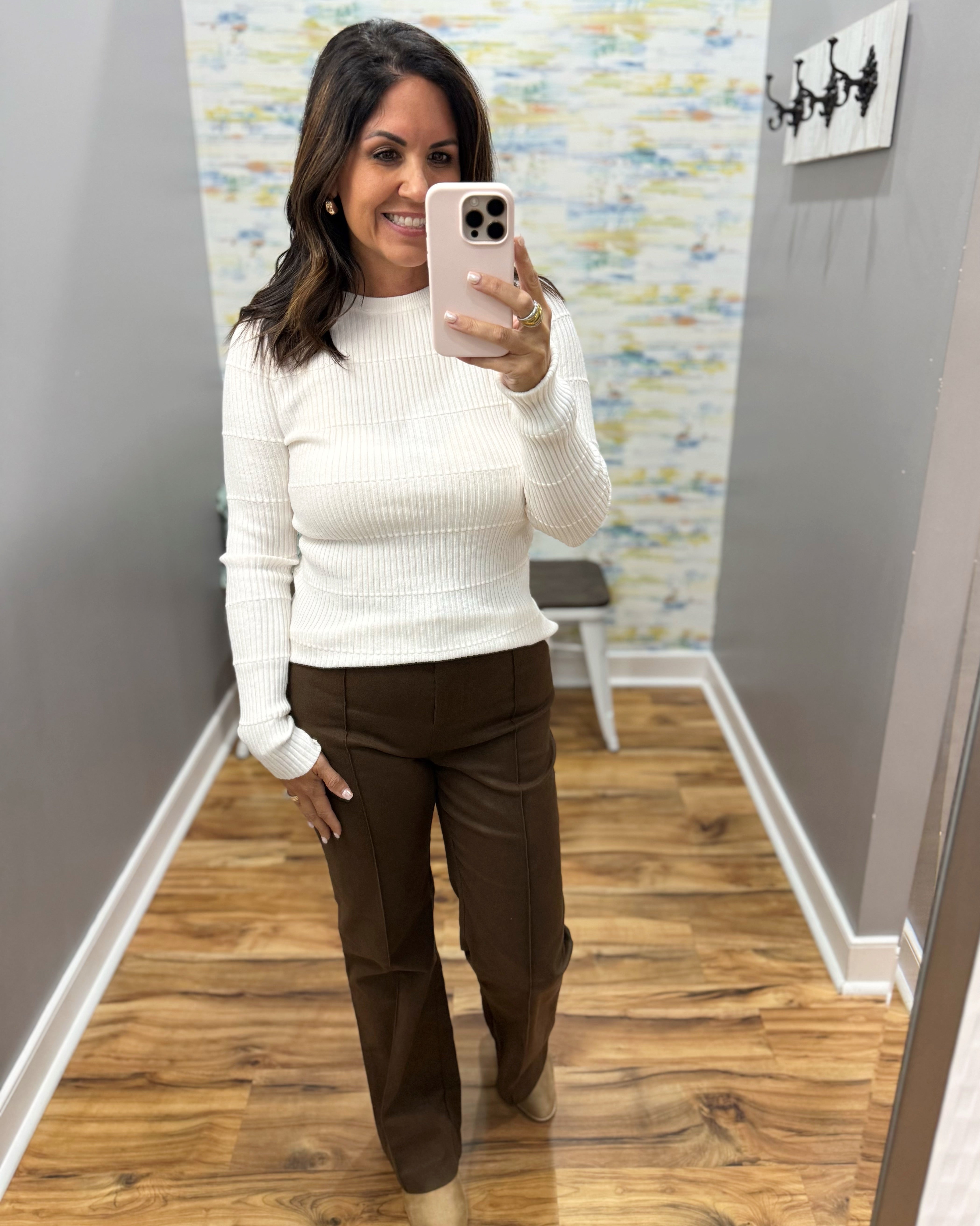 A woman takes a mirror selfie in a fitting room or hallway, wearing a white ribbed long-sleeve mock neck sweater and dark brown wide-leg trousers. She is holding a light pink smartphone.
