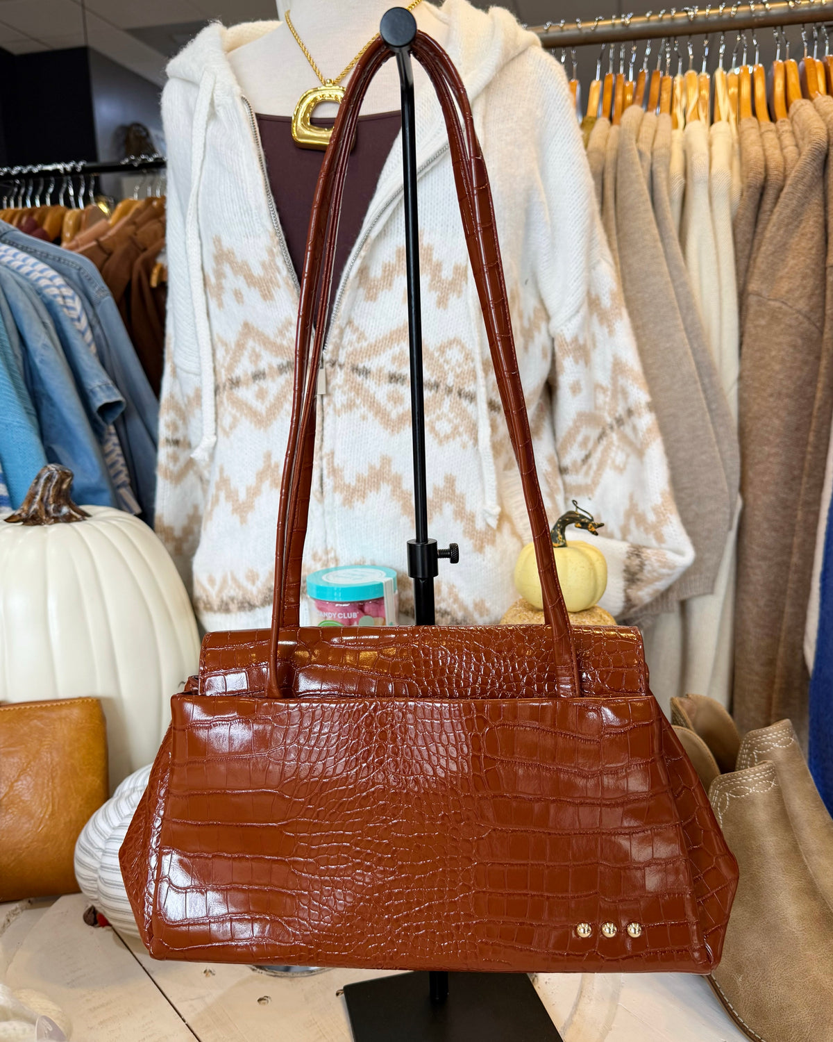 A brown crocodile-embossed leather handbag with double shoulder straps, displayed on a black stand in a clothing boutique setting.