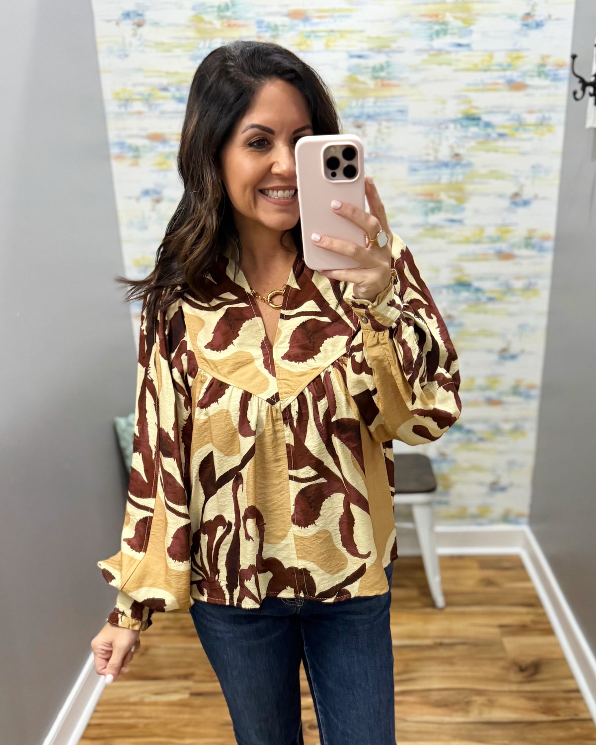 A woman takes a mirror selfie wearing a beige and brown abstract patterned blouse with long ruffled sleeves, paired with dark blue jeans.
