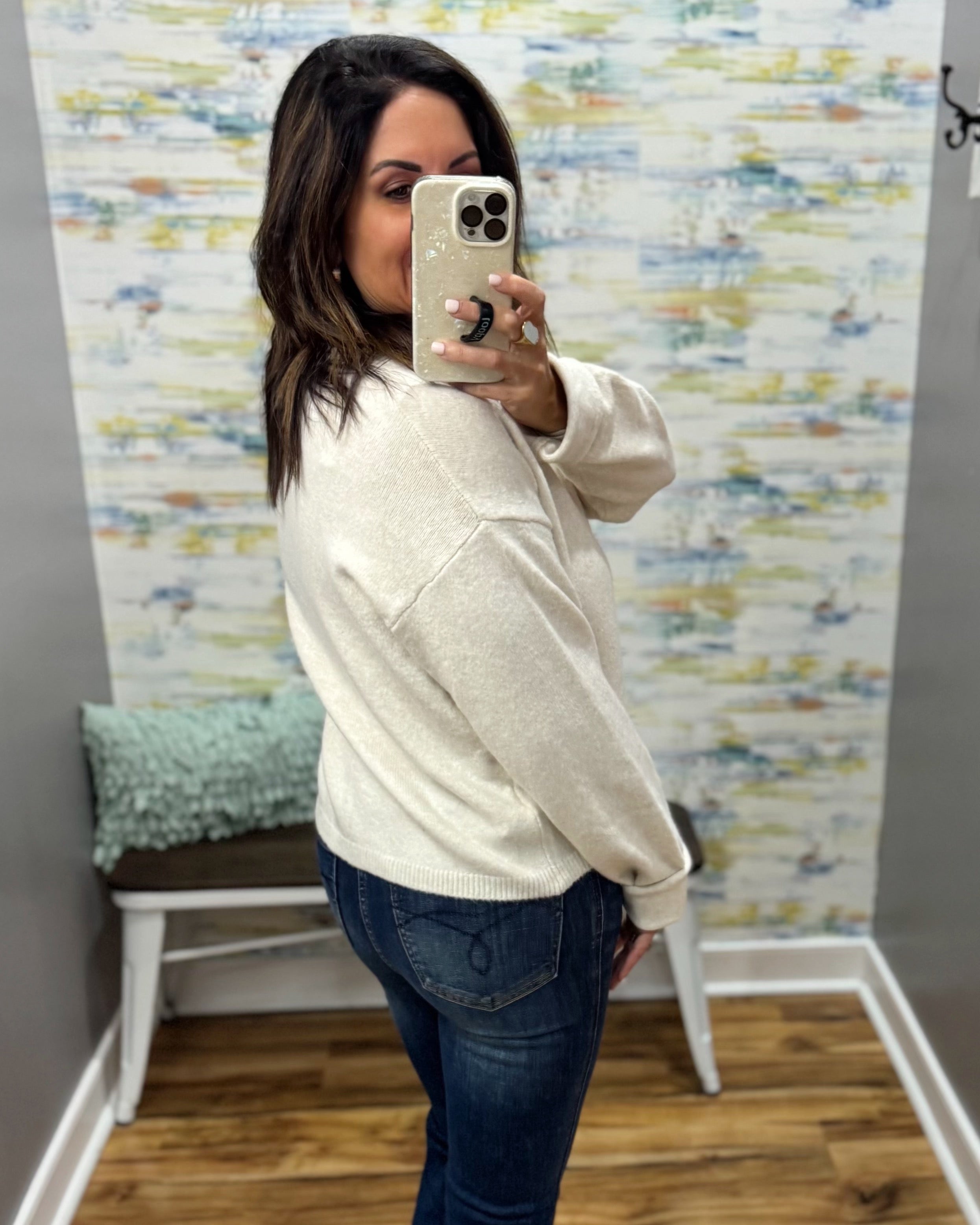 A woman taking a mirror selfie wearing a cream-colored knit sweater and dark blue jeans in front of a patterned wallpaper background.