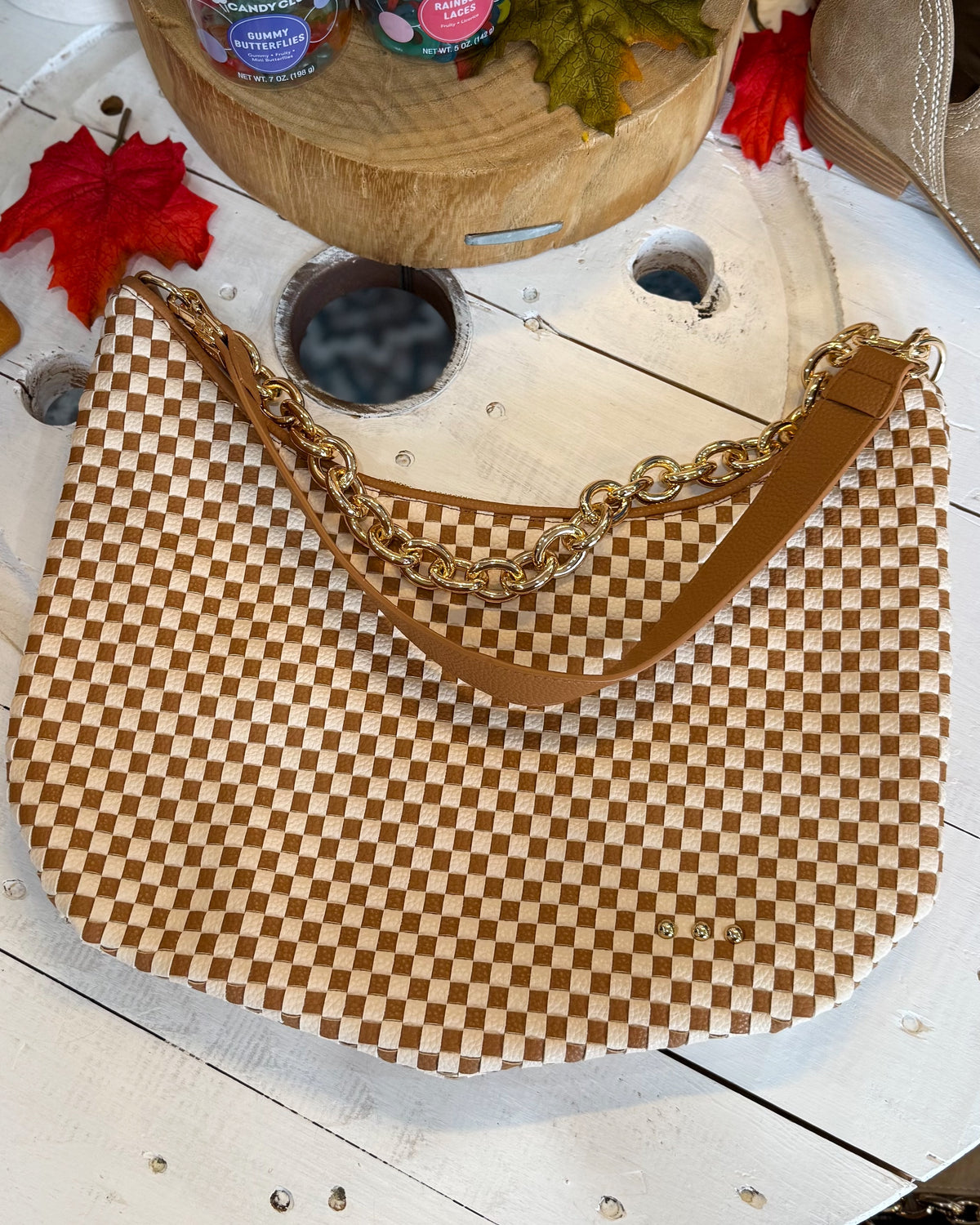 A brown and beige checkered shoulder bag with a gold chain strap, displayed on a white wooden surface with autumn-themed props.