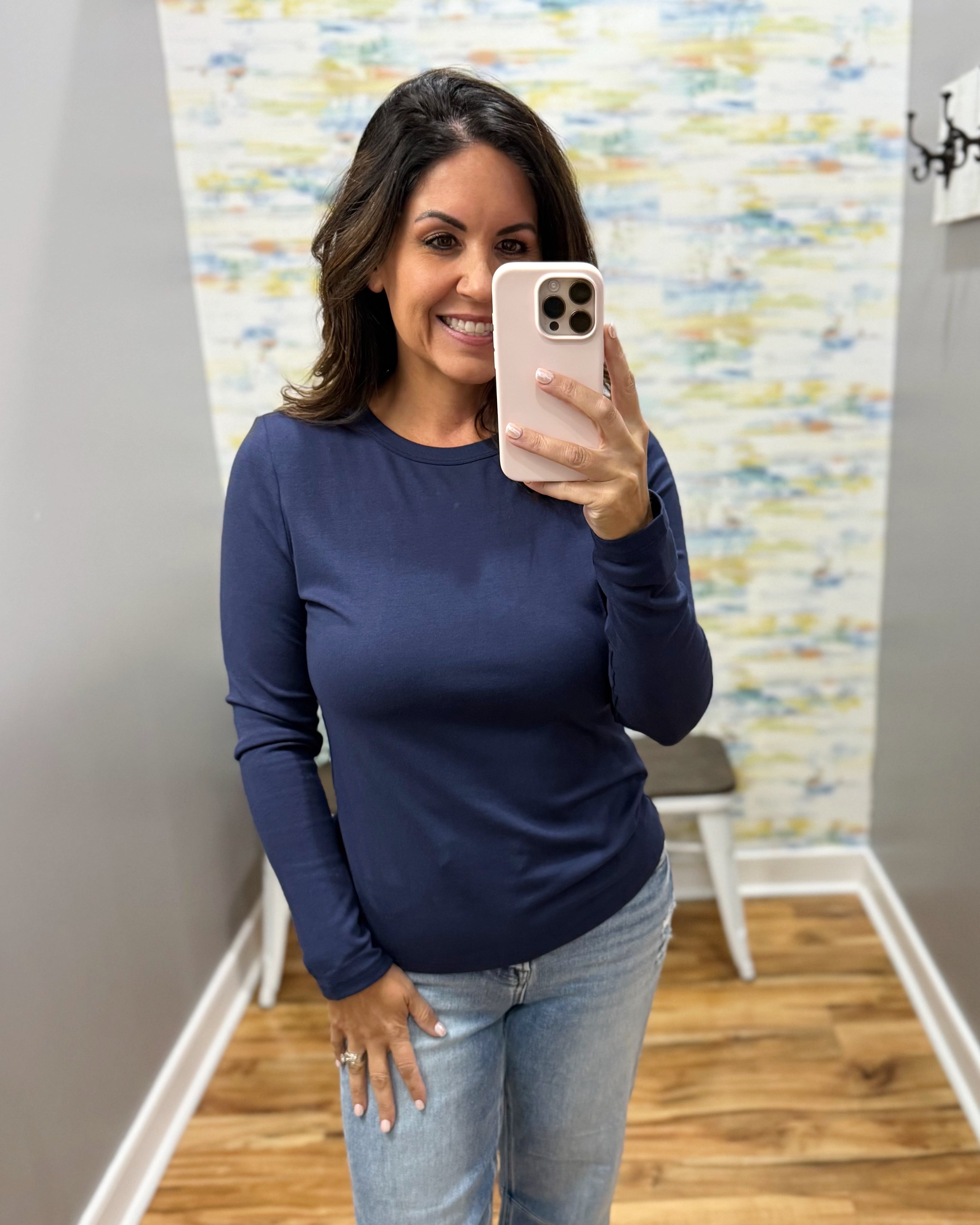 A woman taking a mirror selfie wearing a navy blue long-sleeve crewneck shirt and light wash jeans, holding a light pink smartphone.